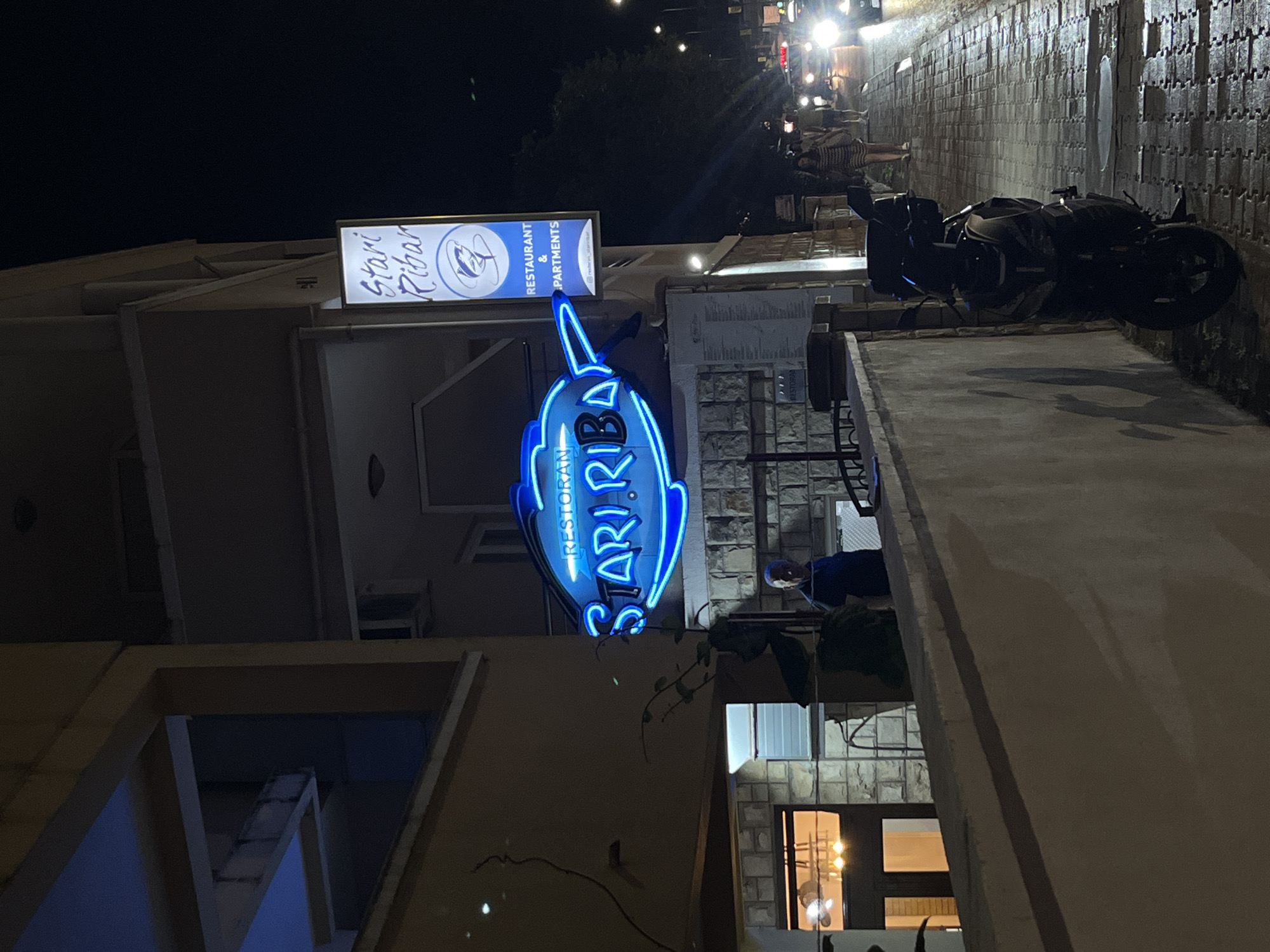 A nighttime exterior shot shows a neon sign in the shape of a fish with the words "RESTORAN STARI RIBAR" in blue neon. To the right, a vertical sign reads "Stari Ribar" in stylized script, with "RESTAURANT & APARTMENTS" below it. A person in a blue shirt stands on a stone-paved area near the restaurant. A parked motorcycle is visible in the foreground. A street with glowing headlights and people walking on a wet sidewalk is in the background, suggesting a city environment. The overall scene is lit by artificial lights and the neon signage.