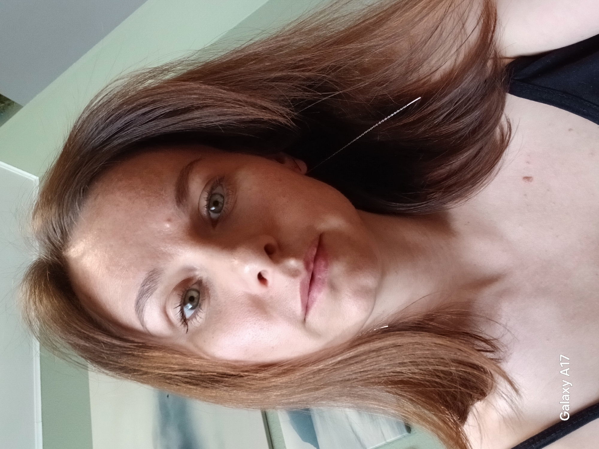 A close-up, angled selfie depicts a woman with light skin, hazel eyes, and light brown hair. She has visible freckles on her face. She is wearing a black top. A delicate silver chain earring is visible dangling from her ear. The background shows a light-colored wall and a partially visible object resembling a painting or picture frame. The phone's watermark "Galaxy A17" is visible in the lower right corner of the image. The image does not contain any information about Yekaterinburg, Russia.