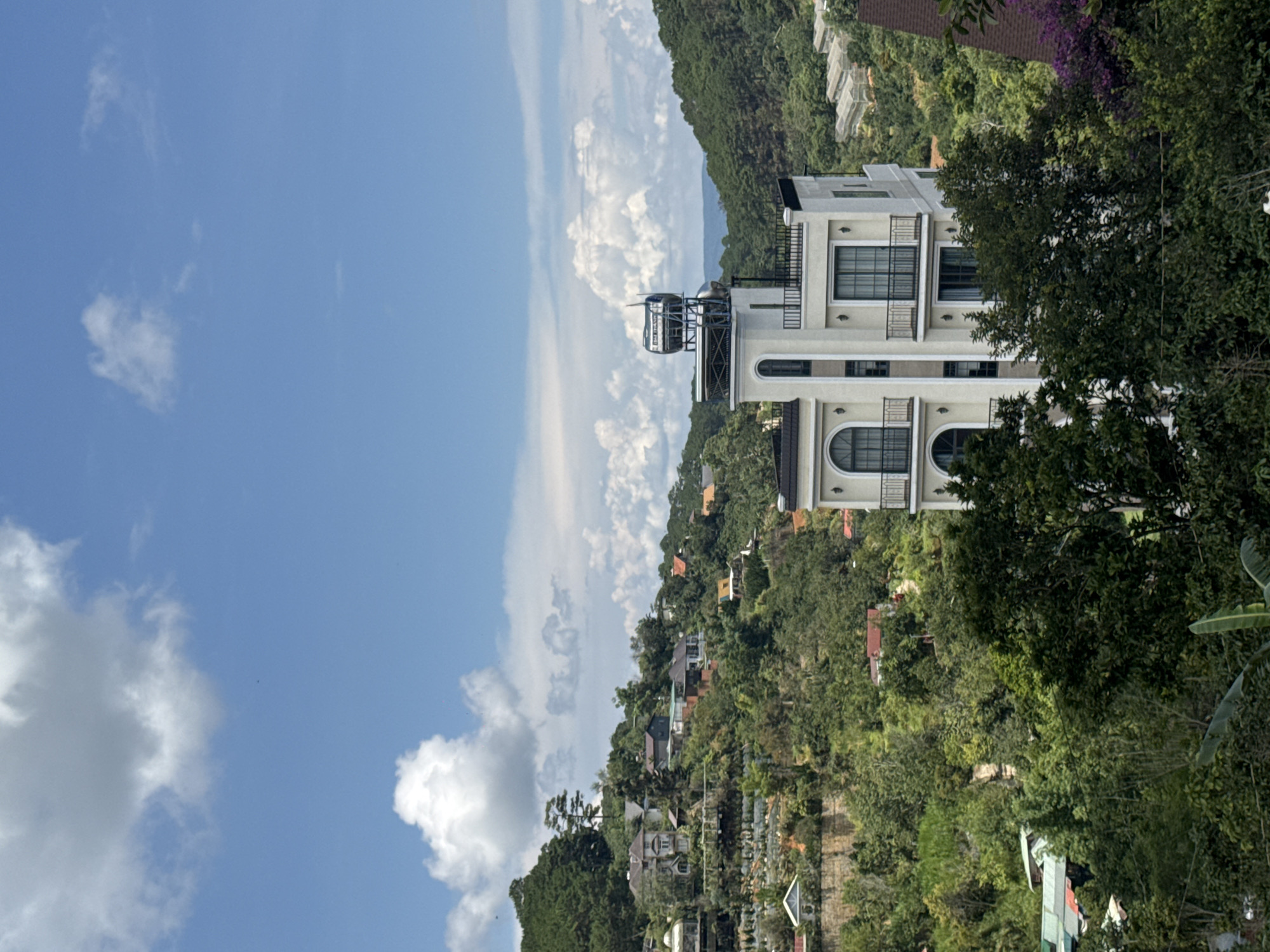 This image captures a scenic view of Da Lat, Vietnam, showcasing a blend of architecture and lush greenery. The foreground is dominated by dense trees, obscuring parts of the lower frame and creating a natural frame for the scene beyond. In the mid-ground, a striking white building with a modern design stands out. It features multiple balconies with black railings, arched windows, and a flat roof topped with what appears to be a solar water heater system. This building is situated on a hillside, surrounded by verdant trees and other smaller structures.
The background reveals a hillside densely populated with houses and buildings, characteristic of Da Lat's hilly terrain. These houses are varied in architectural style and color, nestled amongst the trees, creating a charming, picturesque landscape. Further in the distance, rolling hills covered in pine trees recede into the horizon. The sky is a clear blue, punctuated by fluffy white clouds, suggesting a pleasant day. The overall impression is one of a tranquil, picturesque town set against a backdrop of natural beauty.