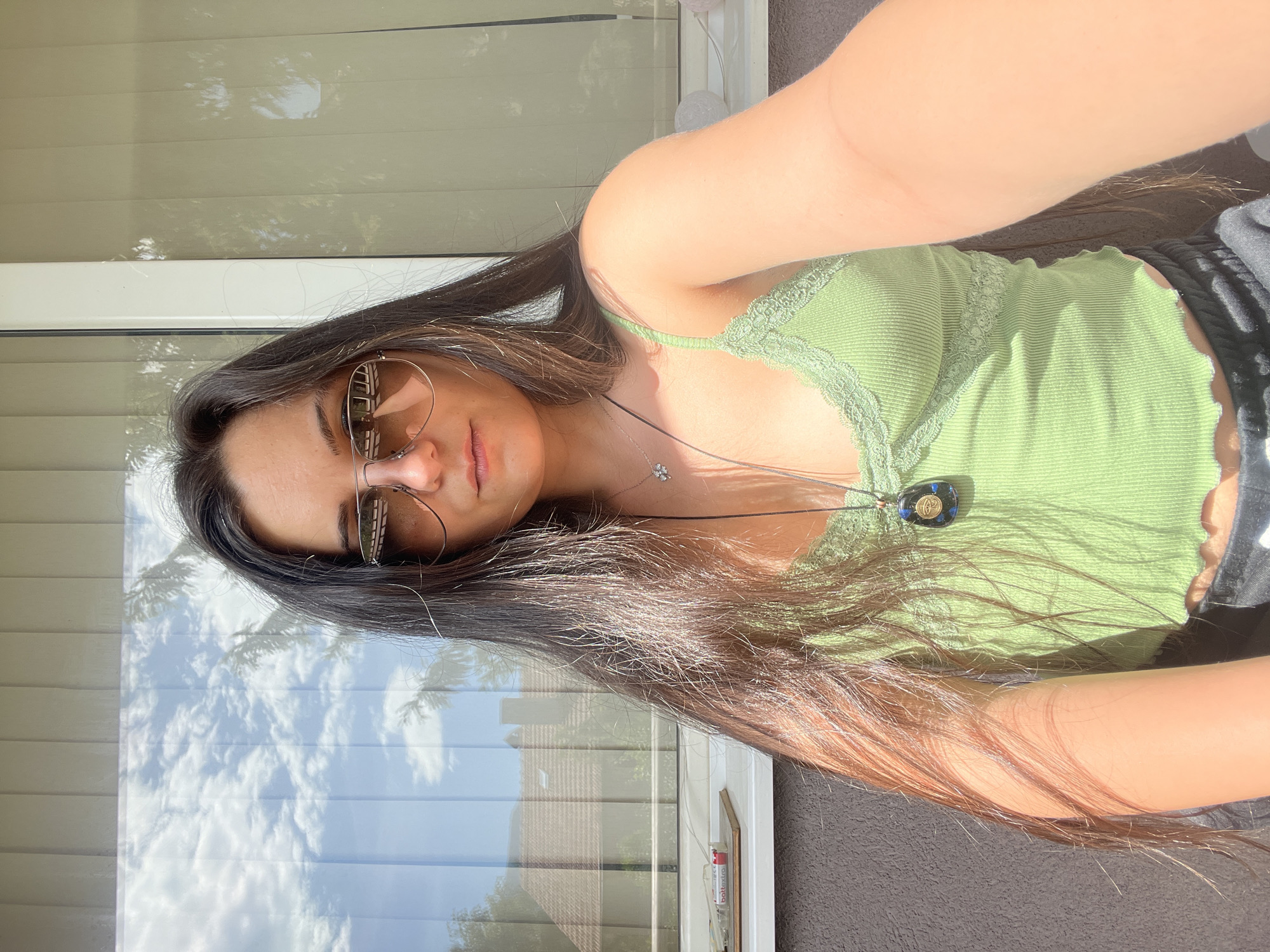 A young woman with long, dark brown hair is taking a selfie outdoors. She is wearing aviator sunglasses, a green tank top with lace trim, and dark pants. She is wearing two necklaces, one with a small floral pendant and another with a large, dark pendant with a blue and gold design. The background shows a building with blinds, and some trees and houses are visible through a window. The lighting suggests it is a sunny day. The location is specified as Sofia, Bulgaria.