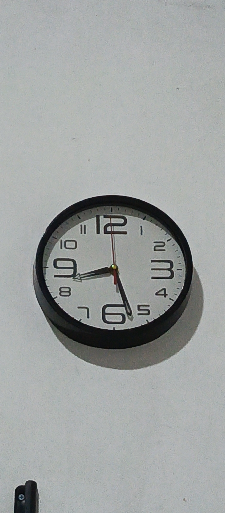 A black-framed analog clock hangs on a white wall. The clock face is white with black Arabic numerals. The hour hand is black and points to approximately 8:50. The minute hand is red and points to 10. The second hand is red and points to 12. The clock is slightly tilted to the left. There is a shadow cast by the clock on the wall below it. In the lower left corner of the image, a small portion of a black electrical device with a silver screw is visible. The wall appears to be painted plaster. The scene is indoors. The time displayed on the clock is 8:50. This object is not associated with a specific location in Sidoarjo, Indonesia.