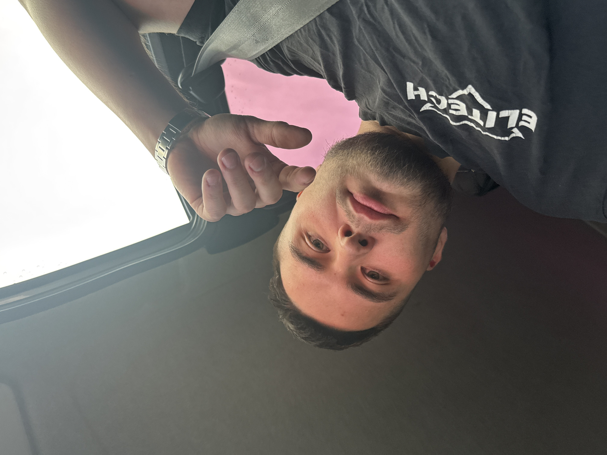 A male individual with short dark hair, a short beard, and light-colored eyes is depicted in a close-up, low-angle selfie. He is wearing a black t-shirt with a white, stylized "ELITECH" logo visible on the left chest area. A silver-toned metal wristwatch is present on his left wrist. His right hand is positioned near his face, with fingers partially curled. A dark grey seatbelt crosses diagonally across his chest. The background indicates the interior of a vehicle, with a dark grey ceiling visible at the top and a car window frame on the right side. Bright white light emanates from a window on the right, and a distinct pink glow is visible through a window or reflective surface behind the subject's left arm.