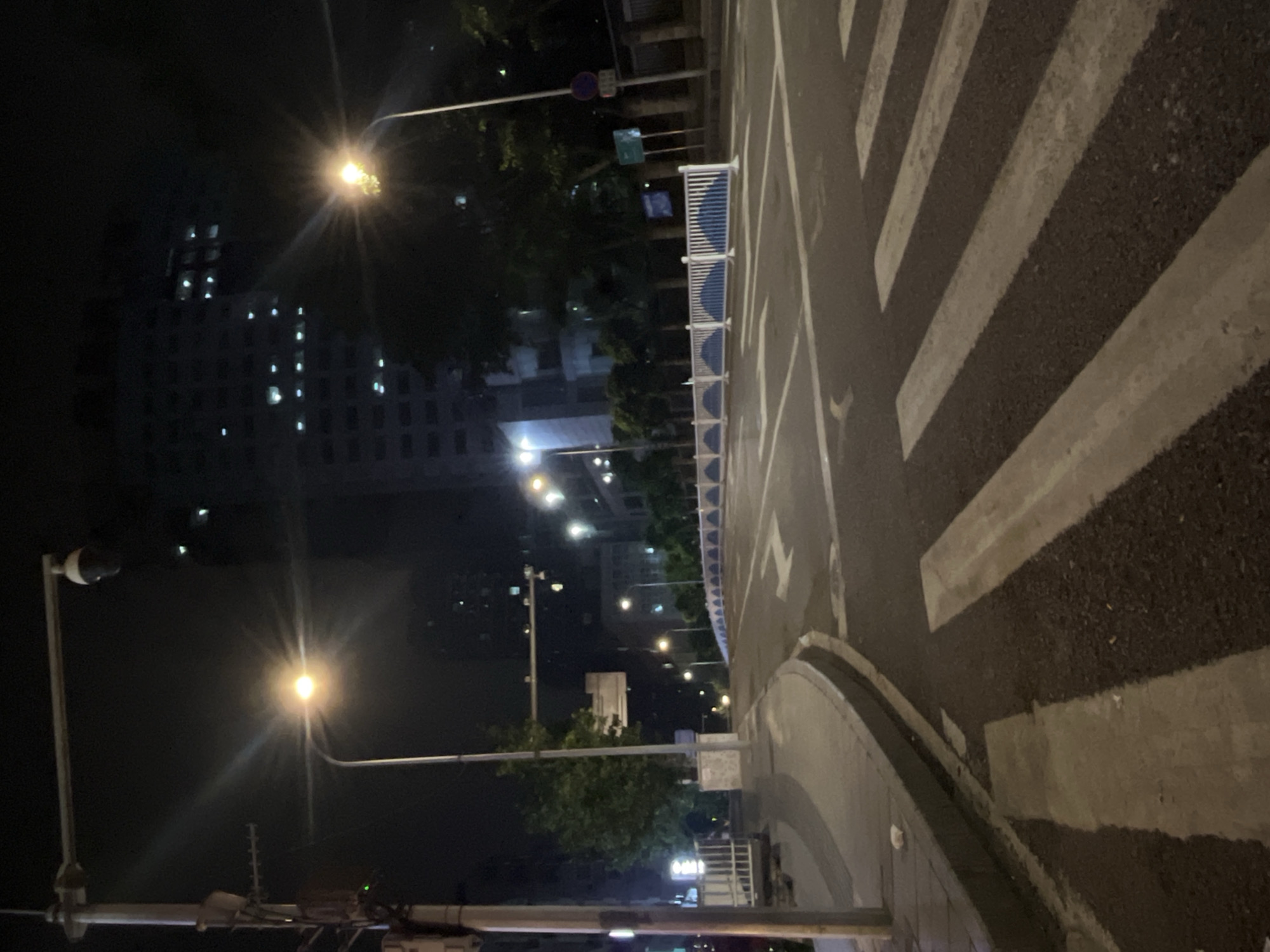An empty, multi-lane street at night in Jiulongpo District, China, curves into the distance. Streetlights illuminate the asphalt, casting long shadows and creating lens flares. A building with illuminated windows rises in the background, partially obscured by trees. Pedestrian crossings are clearly marked with white stripes on the dark road. A white and blue barricade runs along the right side of the street. A lamppost with a camera mounted on top stands on the left. The scene is devoid of any people or vehicles.