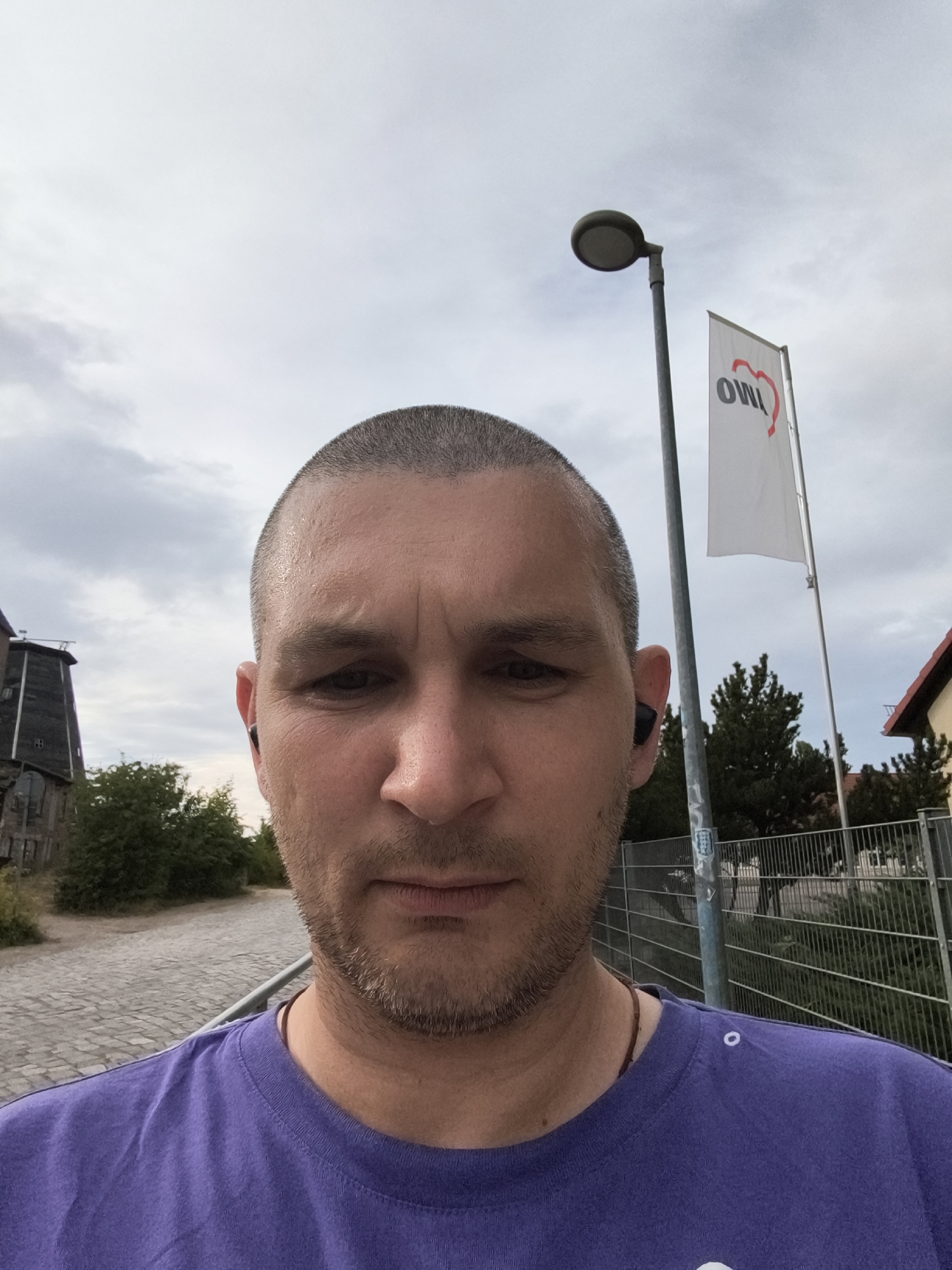 A man with a shaved head and a short beard, wearing a purple t-shirt and earbuds, stands outdoors. He is facing the camera, with a serious expression. In the background, a white flag with red lettering and a heart logo is visible, along with a lamppost. To the left, a dark, industrial-looking building with a tall tower is present. The ground is paved with cobblestones. The sky is overcast. The location is identified as Wolgast, Germany, specifically at the OWiM Museum, a former industrial site turned open-air museum, featuring historical industrial buildings and artifacts.