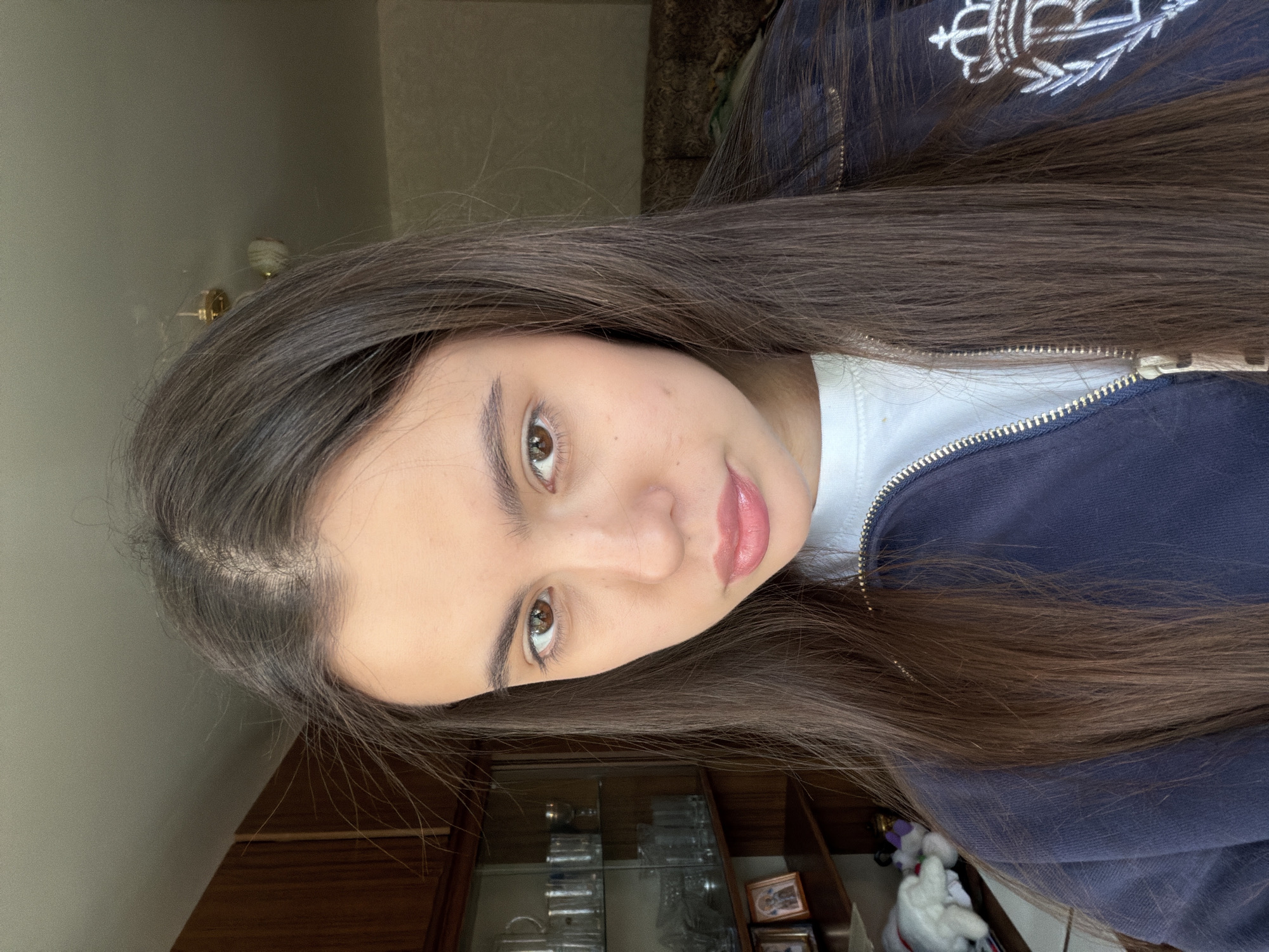 A medium shot of a young woman with long, dark brown hair. She has fair skin and brown eyes. Her lips are pink and appear to have lip gloss on them. She is wearing a white t-shirt and a navy blue jacket with a white embroidered design on the upper left side. The jacket is zipped up partially. The background includes a wall painted in a light neutral color, along with wooden furniture. A display cabinet with glass shelves containing various items can be seen in the lower part of the frame, as well as what seems to be soft toys. The photo was not necessarily taken in the city of Chaikovsky, Russia, as there are no identifiable landmarks within the provided image.