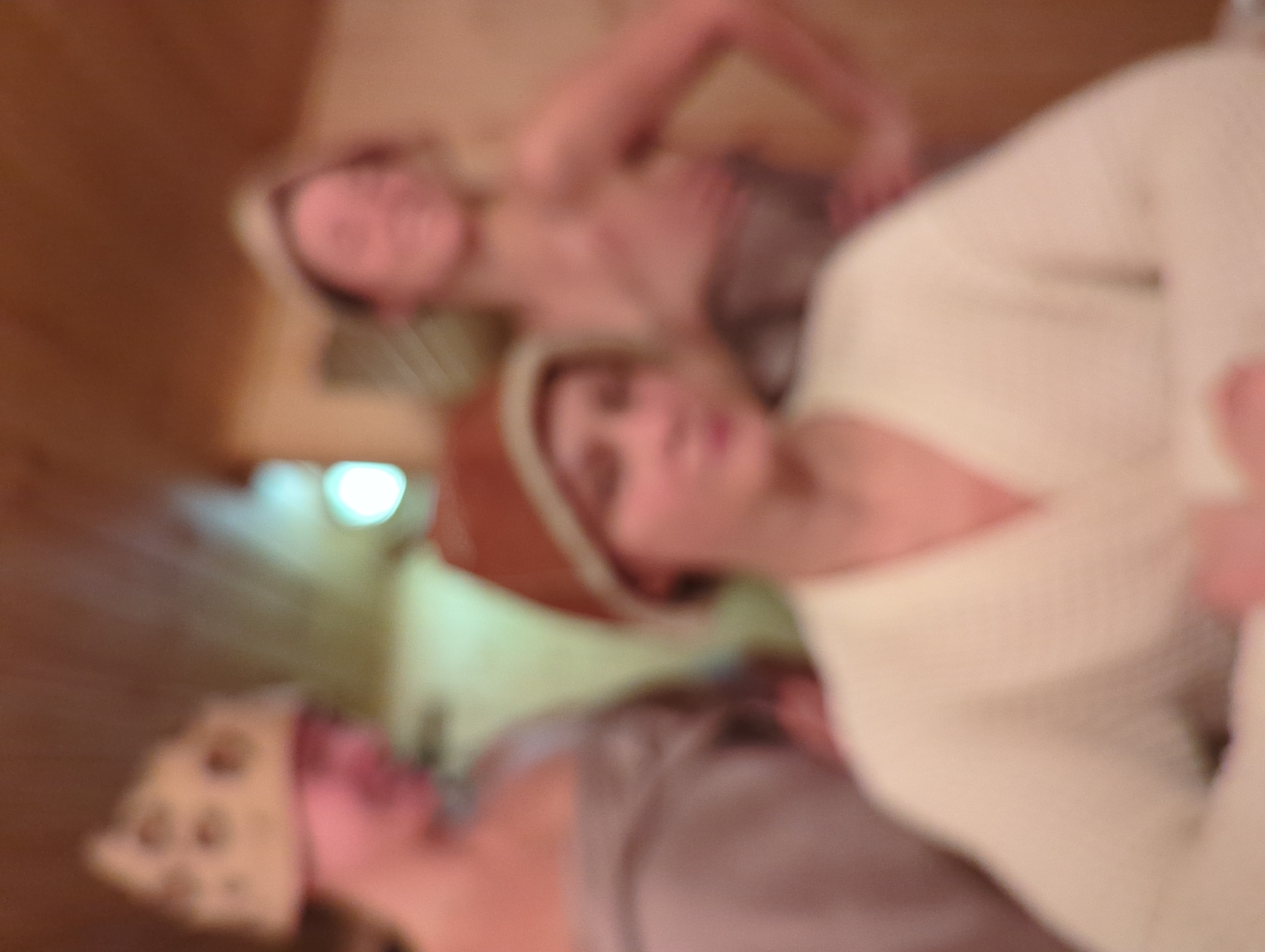 An extremely blurred image depicts three individuals, presumably female, within what appears to be a banya or sauna setting in Khimki, Russia. In the foreground, one individual is partially visible, wearing a light-colored robe and a brown head covering, with hands clasped. Behind and to the left, a second individual is wrapped in a light-colored towel and wears a patterned head covering, facing right. In the background, a third individual stands to the right, also wrapped in a light-colored towel and wearing a cap, with arms bent and head tilted back, suggesting a smile. The environment features light-colored wooden paneling, with a bright light source visible on the left side of the background.