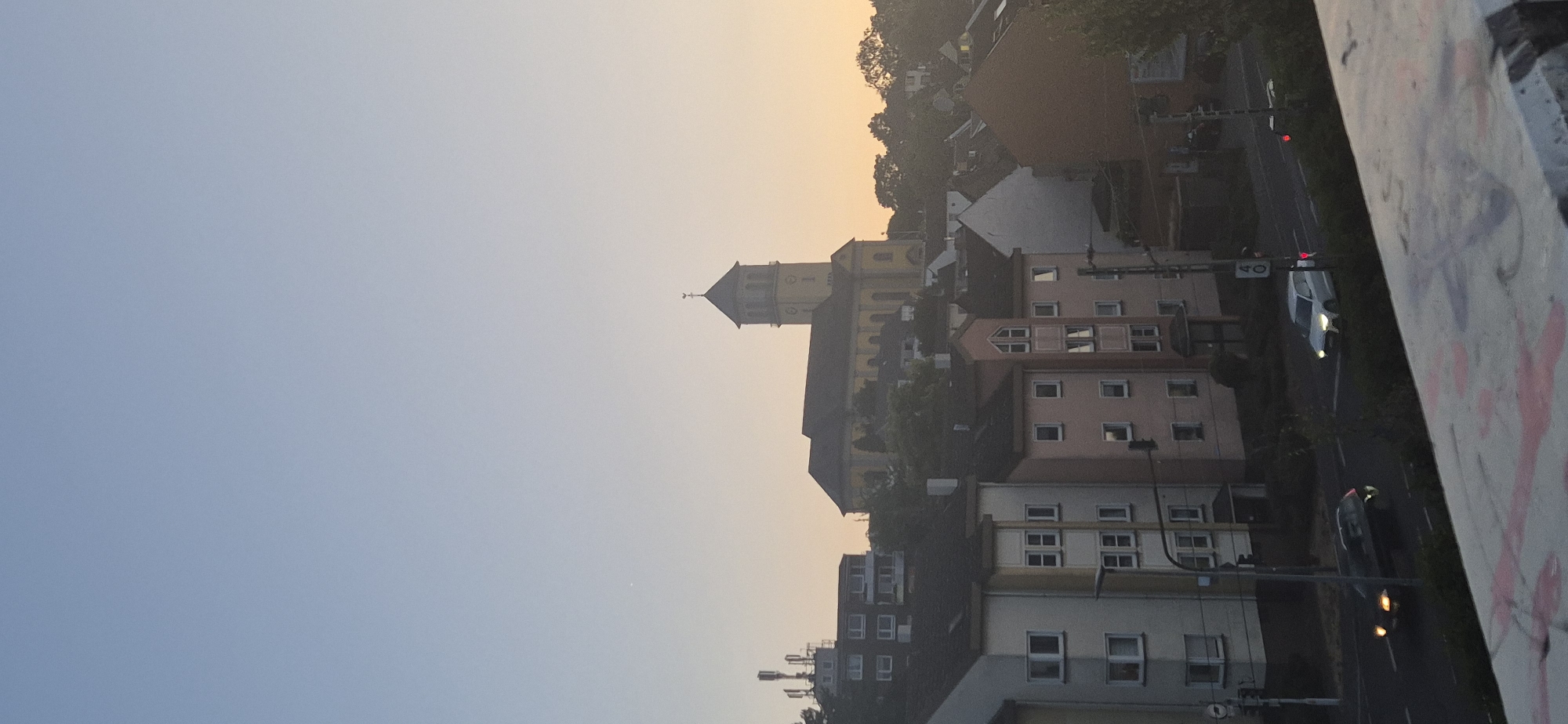 The image captures a view of residential buildings and a church in Mainz, Germany, during what appears to be either sunrise or sunset, given the soft, warm light. In the foreground, a concrete structure with graffiti is visible. Beyond this, a street runs through the scene, with two cars present: a dark sedan on the left and a white SUV on the right. Several multi-story apartment buildings are lined up along the street. In the background, a prominent yellow church with a tall steeple stands on a hill, characteristic of the architecture found in Mainz. The sky above is a pale blue transitioning to a soft orange hue near the horizon, indicating either dawn or dusk. The overall scene depicts a quiet urban environment.