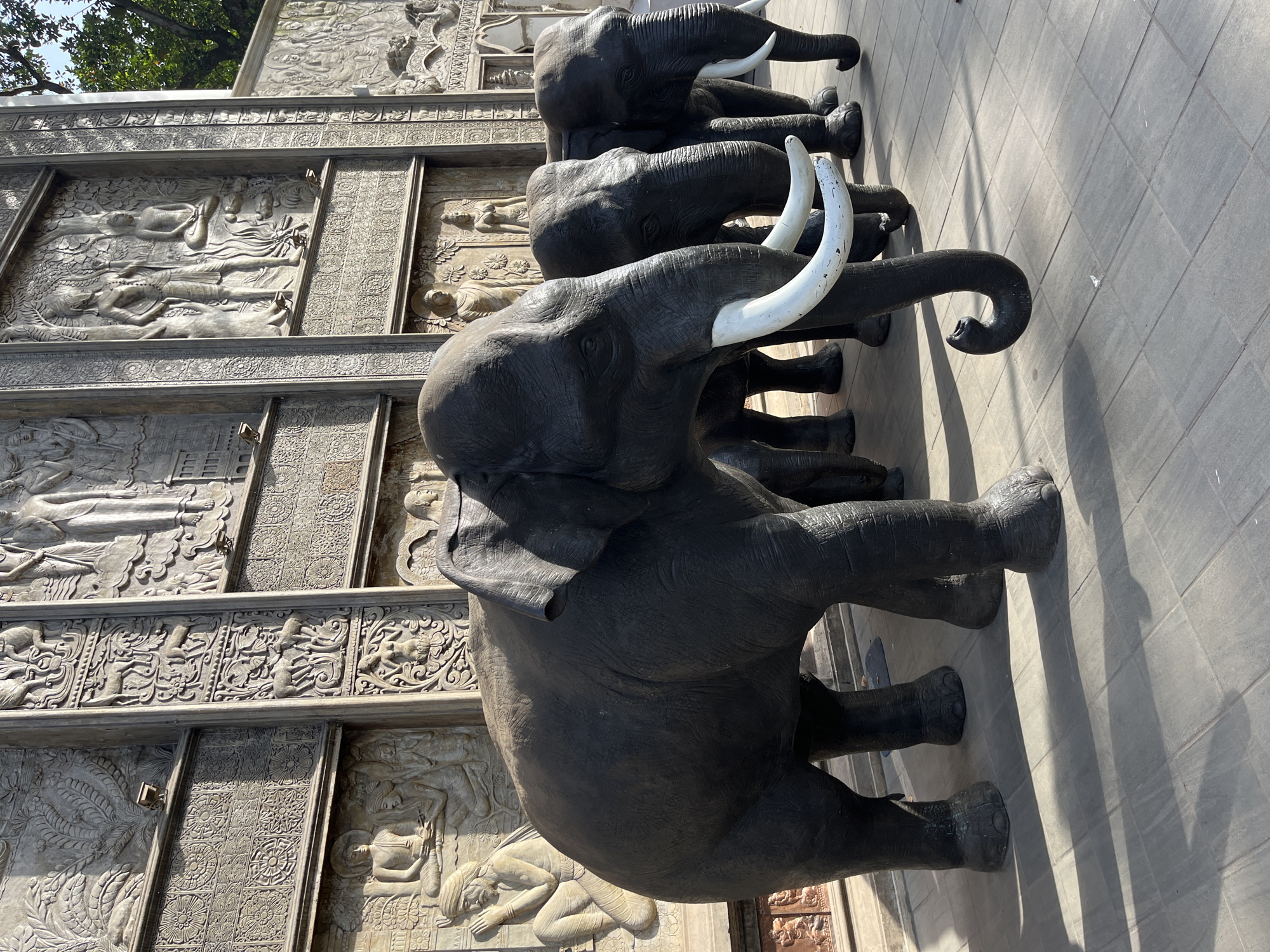The image depicts a row of life-sized elephant statues in front of a building adorned with intricate bas-relief carvings. The statues are dark grey, with prominent white tusks on the two elephants in the foreground. The elephant on the left is the largest and most prominent, with its trunk curled downwards. Behind them, the building features detailed carvings of human figures, animals, and decorative patterns. The scene is set outdoors on a paved walkway, with shadows cast by the statues and the building indicating it is daytime. The overall impression is one of grandeur and cultural significance. This location is identified as the Gangaramaya Temple in Colombo, Sri Lanka.
