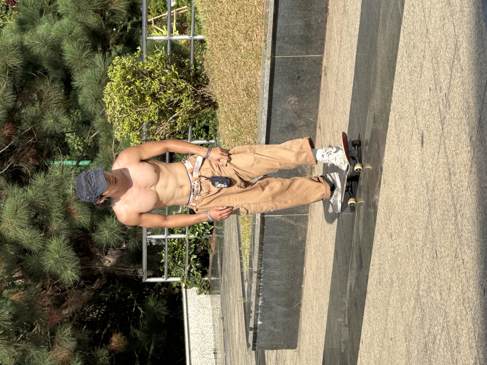 A young man with a lean, muscular physique is skateboarding on a paved pathway. He is shirtless, wearing tan cargo pants rolled up at the ankles, white socks, and athletic shoes. A dark baseball cap is on his head. He is also wearing a belt with a decorative buckle and a bracelet on his left wrist. A small black pouch is attached to his belt.
The scene appears to be outdoors, possibly in a park or public square, with lush green pine trees and bushes in the background. A metal railing runs along a raised planter area filled with dry grass. The ground is a mix of light-colored pavers and a darker, reflective surface that appears wet in places, suggesting recent rain or maintenance. The sunlight casts long shadows across the pavement, indicating it is either morning or late afternoon. This location could be within the grounds of a resort or a well-maintained public space in Da Lat, Vietnam, given the vegetation and manicured environment.