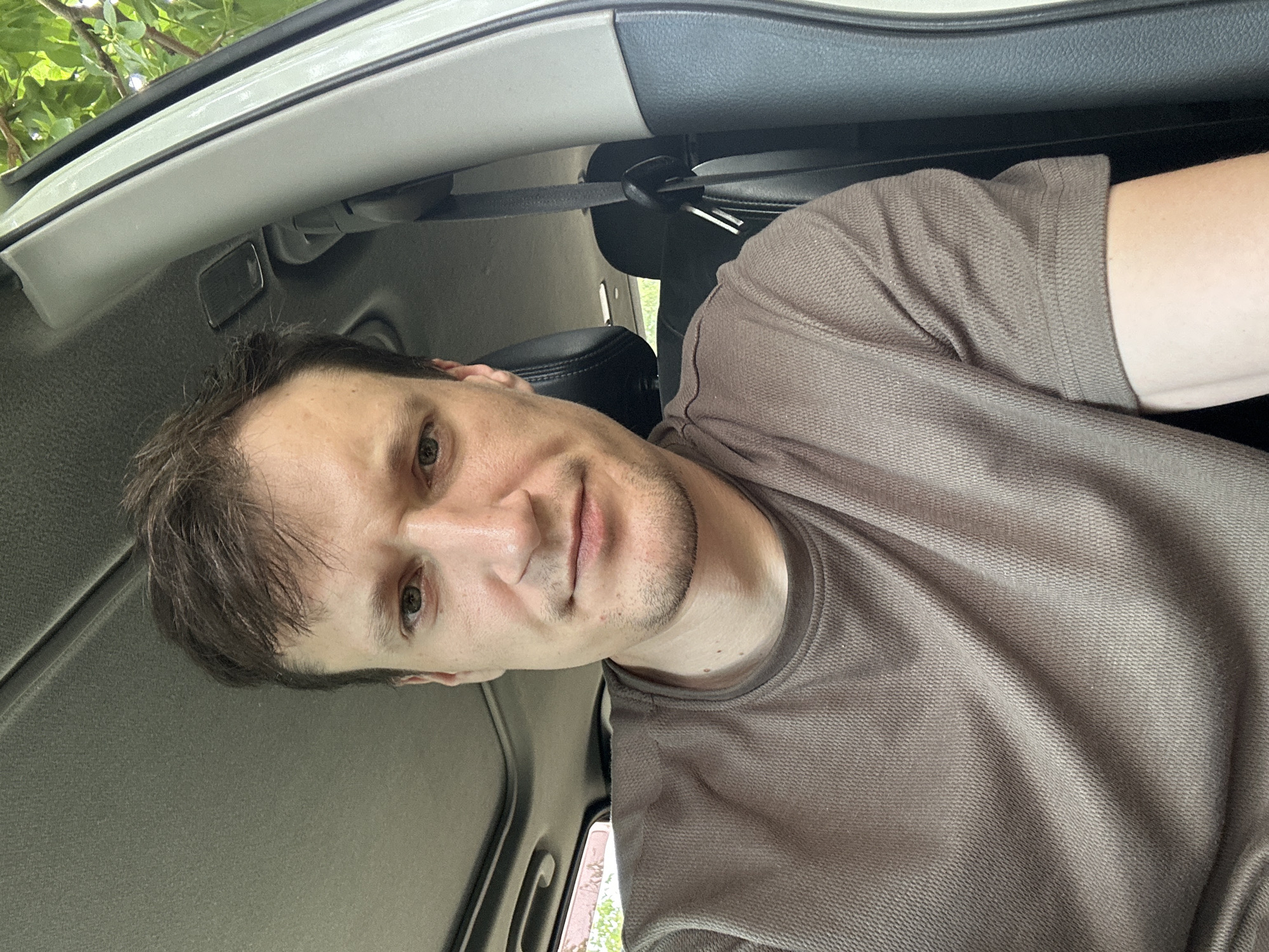 A man is sitting in the driver's seat of a car, looking at the camera. He is wearing a brown t-shirt. The car's interior is visible, including the sunroof and the passenger seat. Outside the car, trees are visible. The image appears to be taken on a sunny day. The location is identified as Makiv, Ukraine.