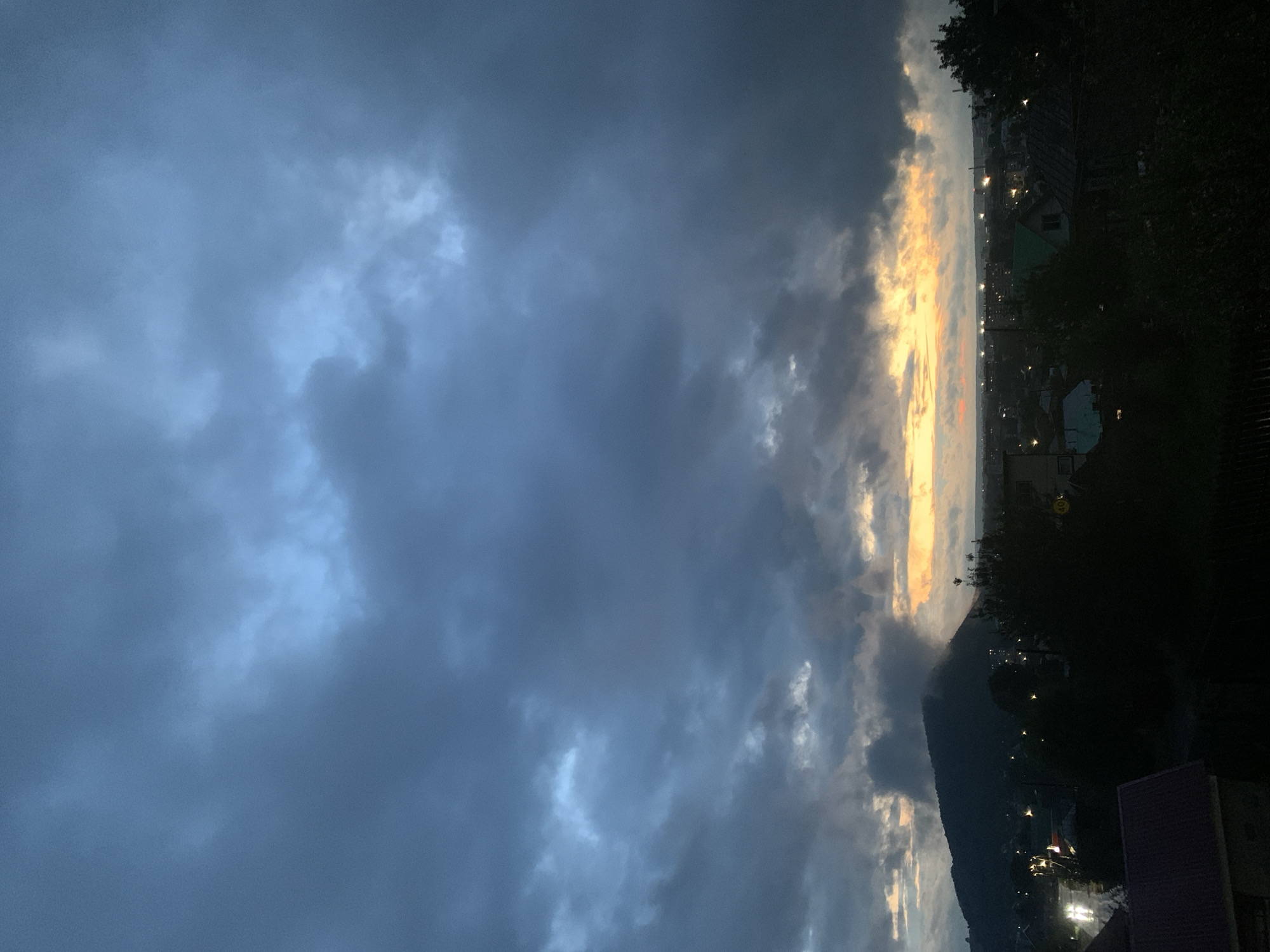 This is a full-frame, high-angle, eye-level shot of a cloudy sunset over the city of Asha, Russia. The sky is filled with dark, moody clouds that are partially illuminated by the setting sun, creating a dramatic contrast of light and shadow. In the foreground, a silhouette of trees and houses creates a dark, ominous atmosphere. A mountain looms in the background, its peak shrouded in mist. The city lights are visible in the distance, twinkling like stars against the twilight sky. The overall impression is one of serene beauty, but also a sense of mystery and foreboding.