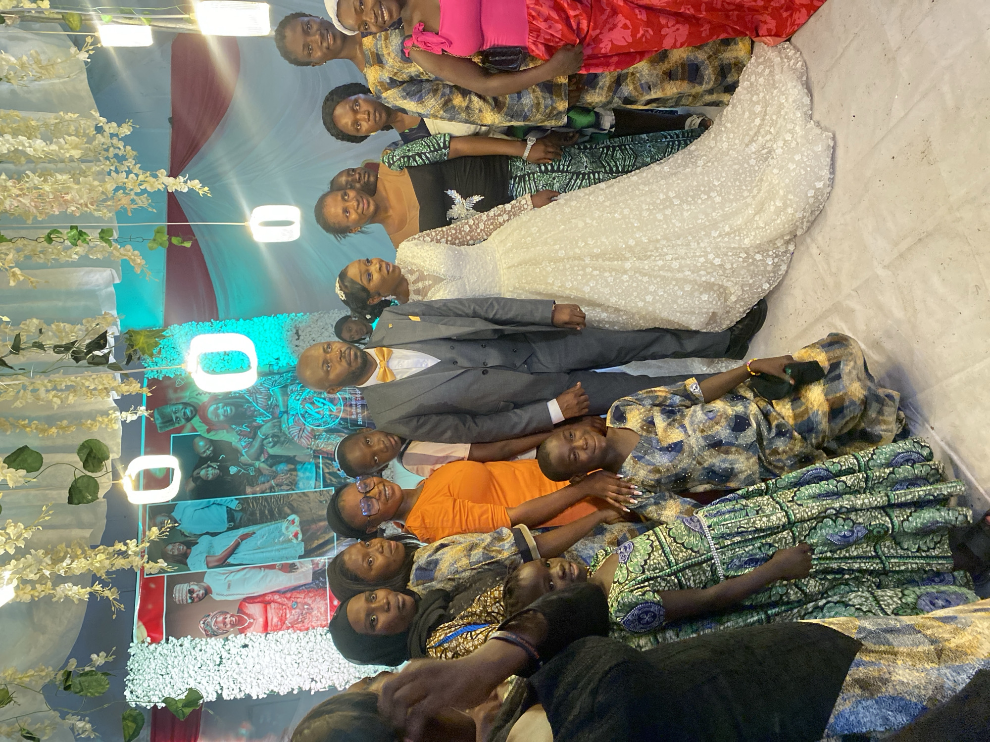 A group of about 15 people are posing for a photograph. The group is vertically oriented in the frame, with a backdrop featuring white flowers, decorative lighting, and portraits of a couple on a patterned surface.
The people in the photo appear to be at a wedding or related event. The group includes a bride in a white lace gown and a groom in a gray suit with a gold bow tie. The other individuals are dressed in a variety of colorful and patterned fabrics. The group is arranged in a line and is mostly smiling.
Based on the faces, people, and dress, the location is Kuru Dungus, Nigeria.