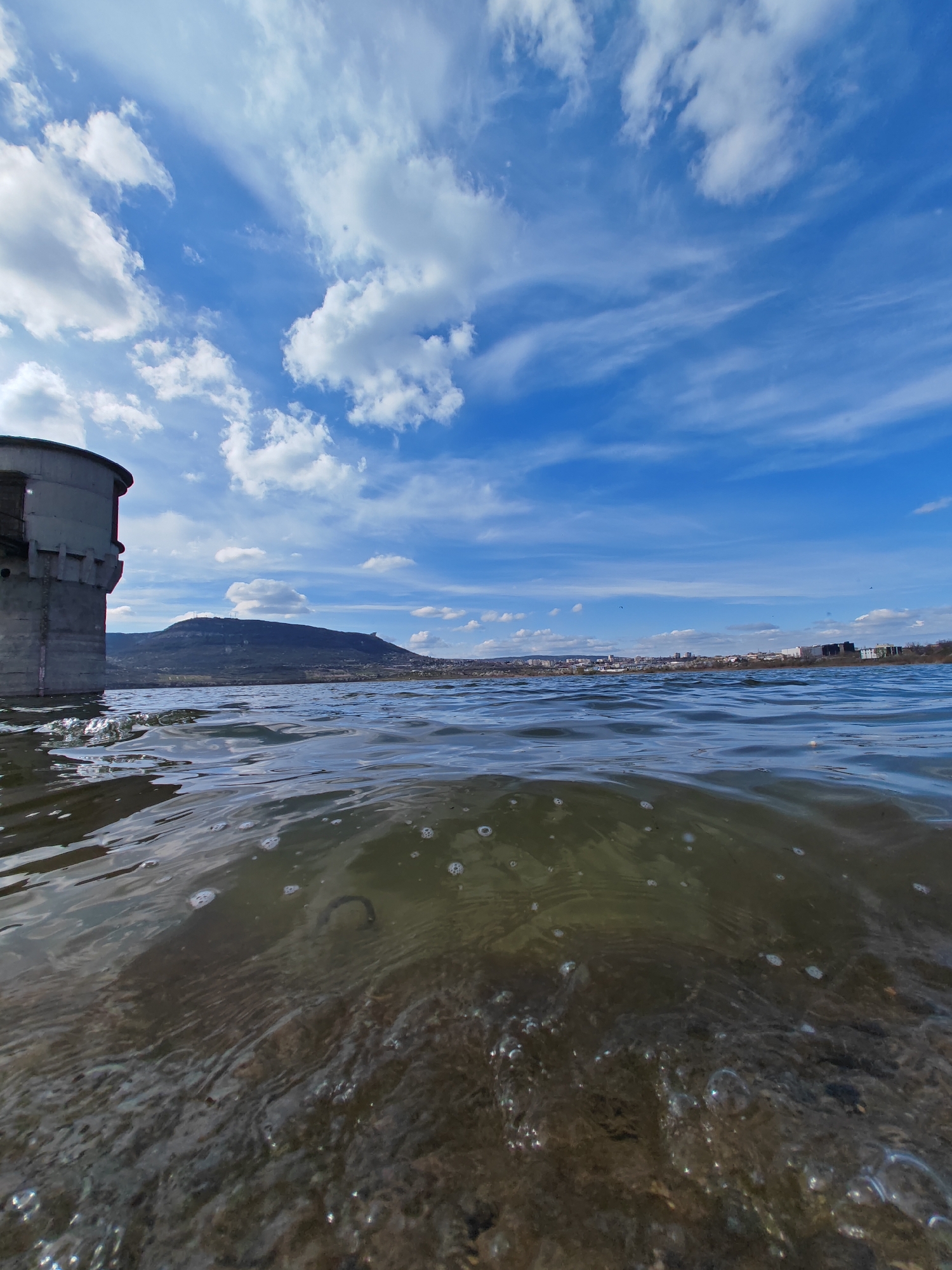A low-angle view, taken at water level, shows a reservoir under a partly cloudy blue sky. In the lower half of the image, water obscures the view of the reservoir bottom, which appears to be stony. Bubbles are visible in the water. The water transitions to waves at the water line, above which is a view of a distant city and a tree-covered hill. To the left, a grey concrete tower structure stands partially submerged in the water. This scene is likely from the water reservoir on the road to the Shumen Tomb.