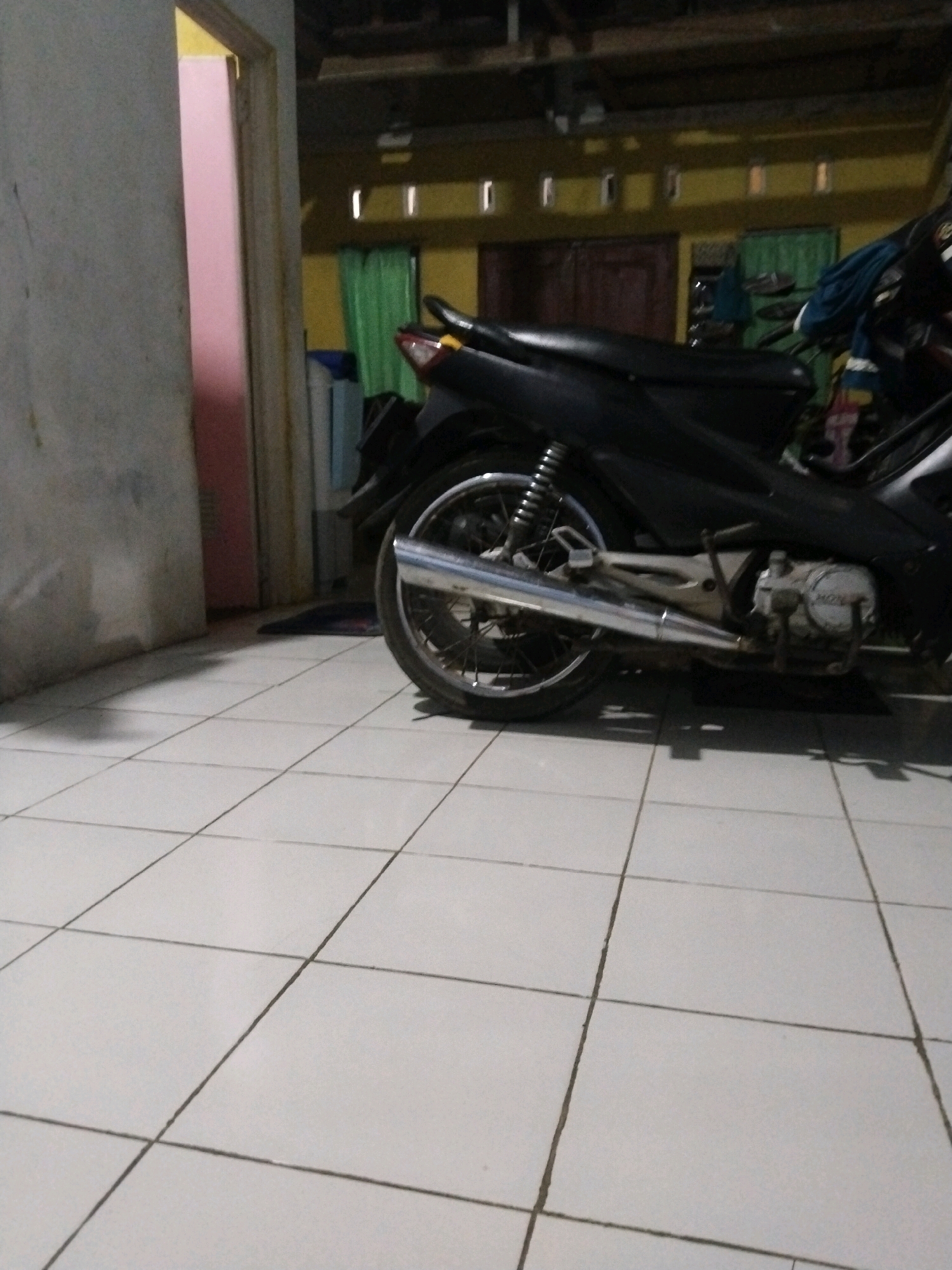 The image depicts an indoor scene, likely a residential area, with a motorcycle parked on a tiled floor. The motorcycle is black and appears to be a scooter-style model. Its rear wheel, exhaust pipe, and engine are visible. To the left of the motorcycle, there is a doorway leading to another room, which appears to be painted a pale pink. The background shows a yellow wall with small rectangular windows, and green curtains hang in front of them. Dark brown wooden doors are also visible in the background. The floor is covered with white square tiles, with darker grout lines. The lighting suggests it might be late afternoon or evening, with some shadows cast on the floor. The overall impression is one of a domestic setting, possibly in a home or a covered parking area within a residential compound in Werasari, Indonesia. There are no people or explicit actions captured in the frame.