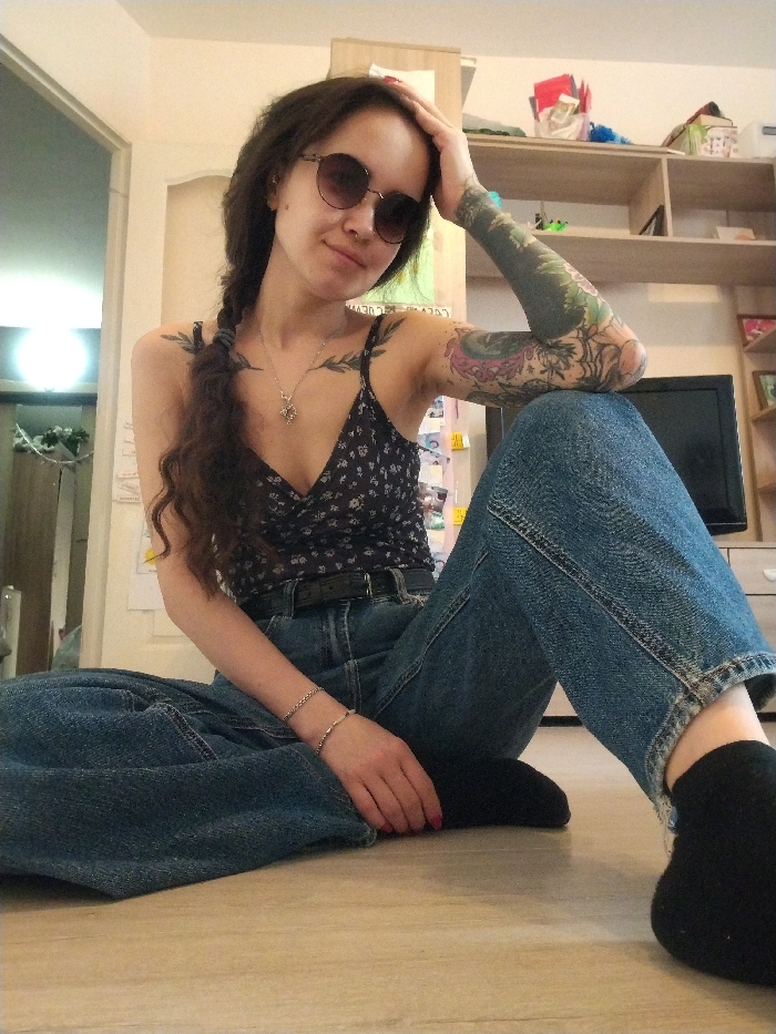 A woman sits cross-legged on the floor wearing a black floral print top, blue jeans, and black socks. She has long dark hair braided to the side, sunglasses, and a tattoo sleeve on her left arm. She rests her left hand on her head. Behind her is a shelf unit with various items and a television. A doorway is visible in the background to the left. The floor is light wood.