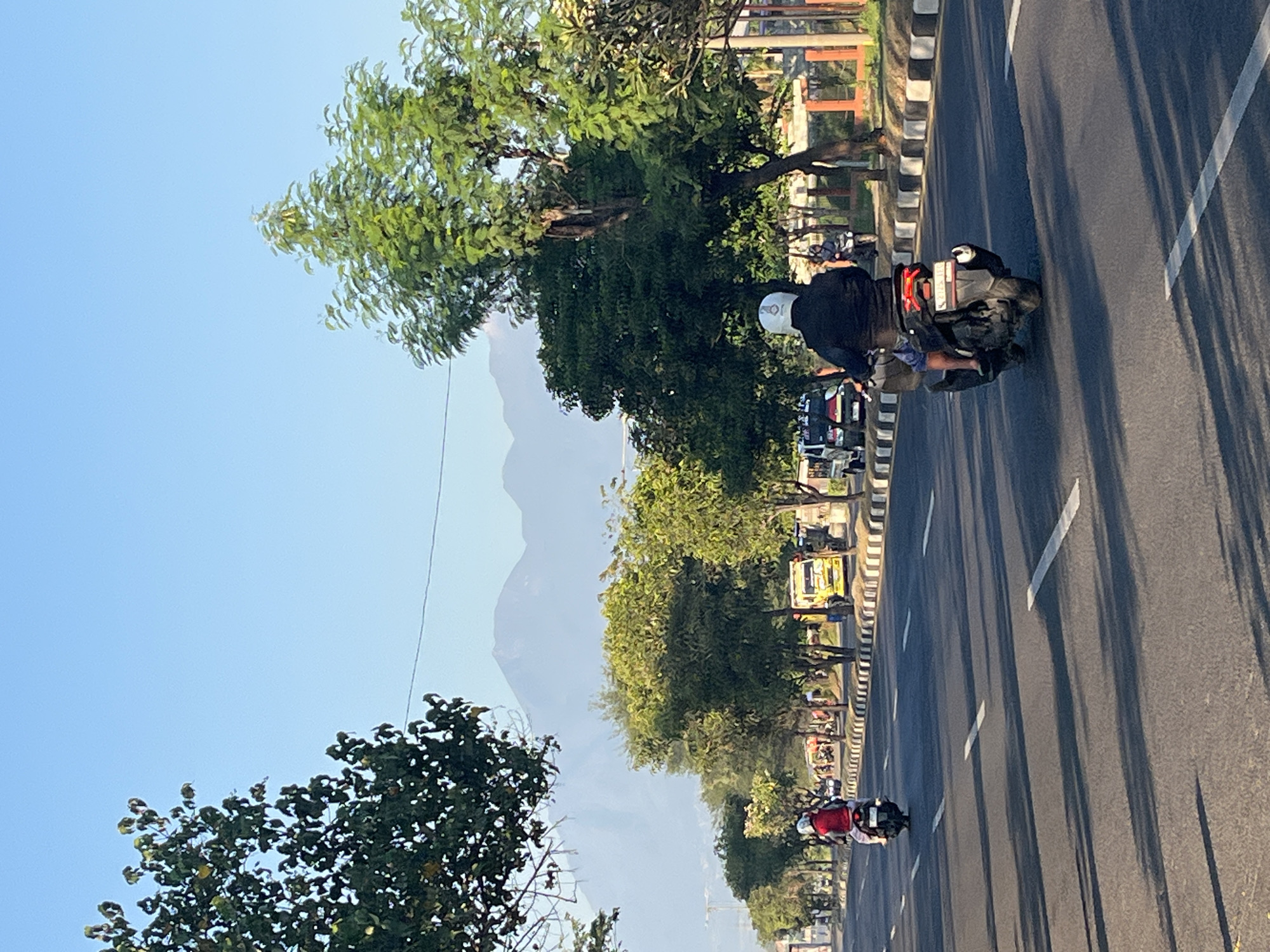 In Siring, Indonesia, this image captures a sunlit street scene with vehicles and lush greenery. Two motorcycles are prominent on the asphalt road. The first, further down the lane, carries a person in a red shirt. Closer to the viewer, a second motorcycle is ridden by an individual wearing a black jacket and a white helmet. In the background, a yellow truck is visible, alongside other vehicles and a row of trees lining the street. Beyond the trees, distant mountains rise against a clear blue sky, hinting at the natural landscape surrounding the urban environment. The road is marked with white lines, and shadows of trees stretch across the pavement, indicating the time of day.
