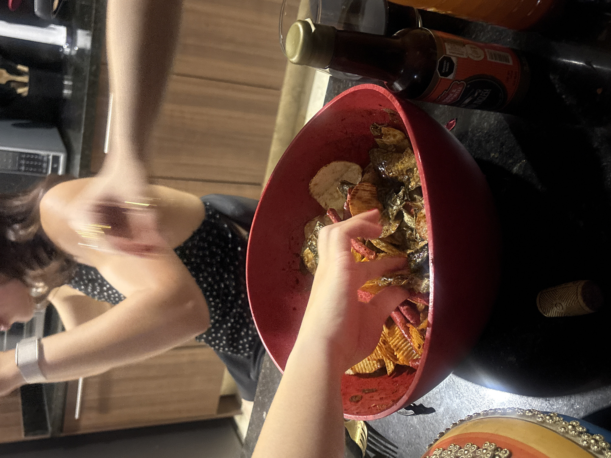 A medium shot captures a red bowl filled with chips and some kind of meat on a black counter. A bottle of salsa inglesa sits beside the bowl. A person's hand reaches into the bowl. Another person wearing a black and white dress is out of focus in the background. A fork is placed near the bottom of the bowl. An item with colors of blue, yellow, and orange is at the lower part of the frame. The location is unknown.