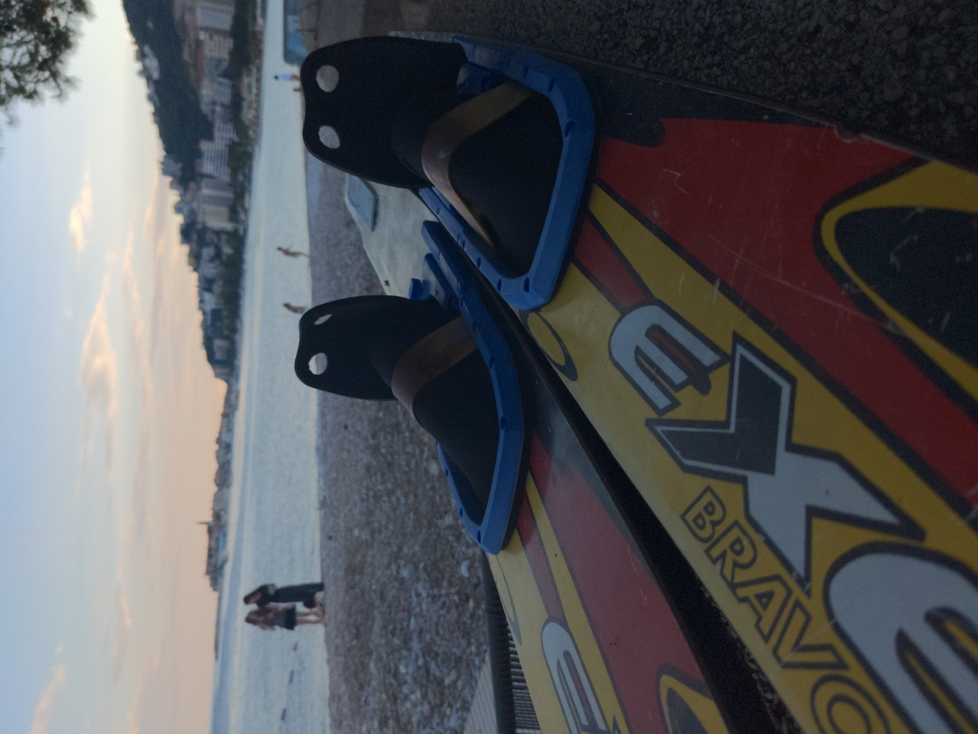 The image shows a pair of water skis lying on a pebble beach. The skis are red, yellow, and black with the word "EX BRAVO" visible. They have blue bindings with black foot holders. In the background, there is a body of water and a coastline with buildings, likely a city. Two people are standing on the beach near the water's edge. The sky is pale. The location is Unknown, Unknown.