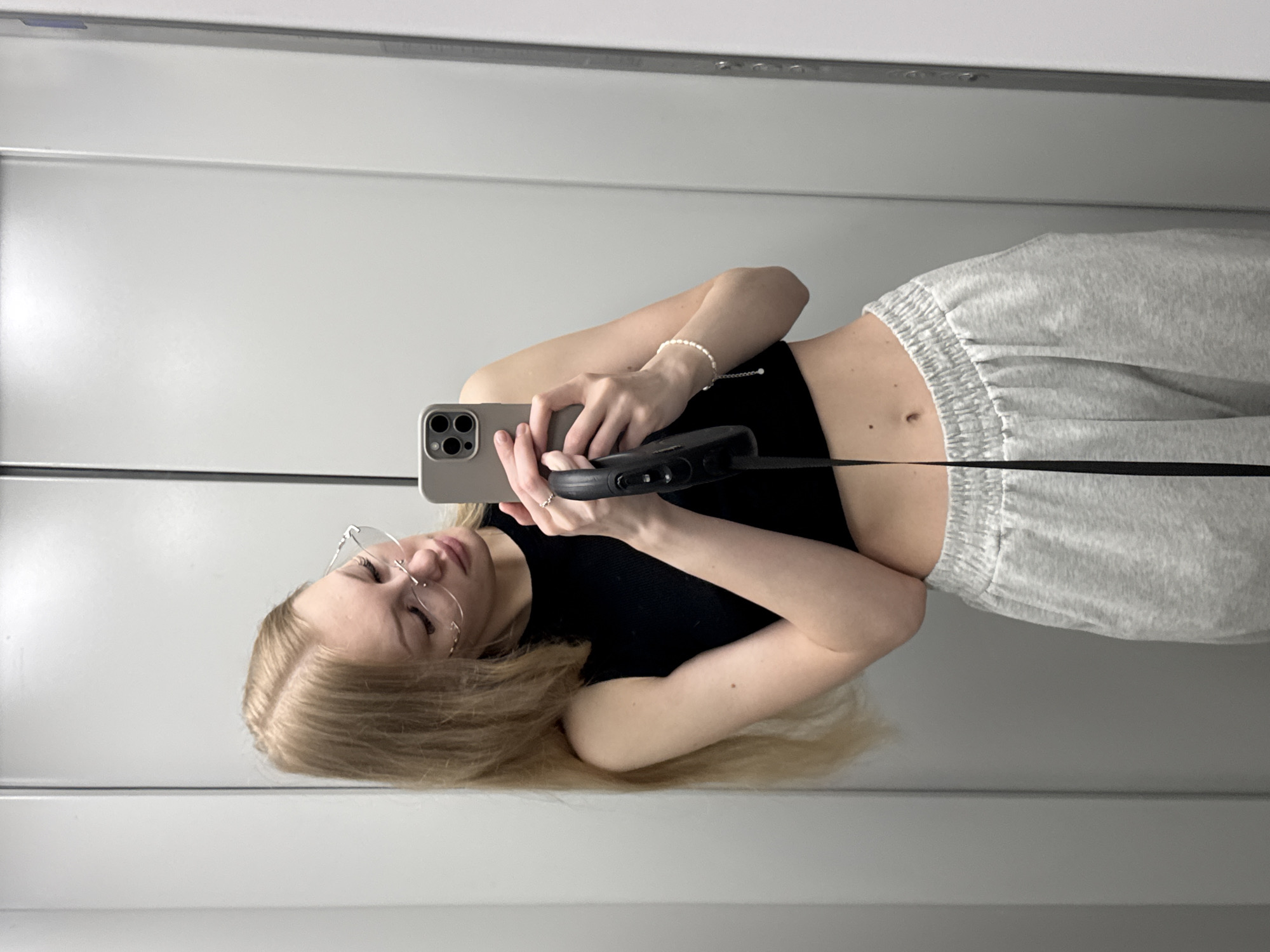 The image is a mirror selfie taken by a young woman with blonde hair and glasses. She is wearing a black crop top and light gray sweatpants. She is holding a smartphone in her right hand, taking the picture. A black strap from the phone case hangs down. A beaded bracelet is visible on her left wrist. The background appears to be an elevator with gray doors. There is no specific indication of the city of Perm, Russia in the image.