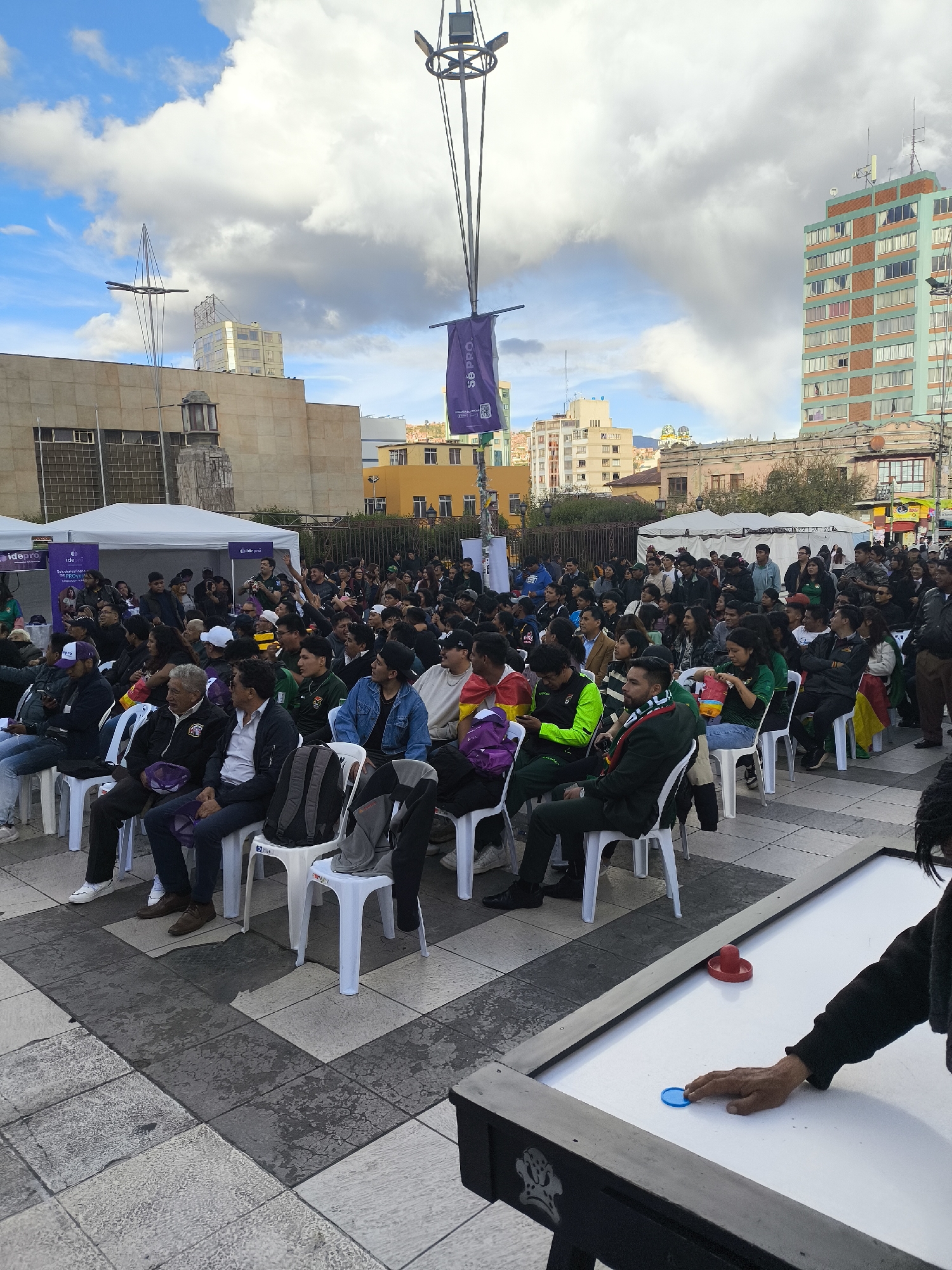 An outdoor public gathering is depicted in Plaza San Francisco, La Paz, Bolivia, under a partly cloudy sky. Approximately 100 to 150 individuals, primarily young to middle-aged adults, are seated in white plastic chairs arranged in rows across a paved plaza, facing an unseen focal point to the left of the frame. Many attendees are wearing casual clothing; some display the national colors of Bolivia (red, yellow, green) on scarves or draped flags. In the immediate foreground, the corner of an air hockey table is visible, where a person's right hand is positioned over a blue puck, a red mallet resting nearby.
In the background to the left, a large, light-colored concrete building with a prominent facade and a distinctive street lamp/monument structure is visible. Further left, two white canopy tents are set up, one bearing the partial branding "Idapro." A tall, ornate metal structure stands centrally, from which a purple banner with "SOBRO" and a QR code is suspended. To the right, various urban buildings rise, including a distinctive multi-story structure with teal-green and reddish-brown sections. The ground is paved with irregular grey and off-white square and rectangular tiles.