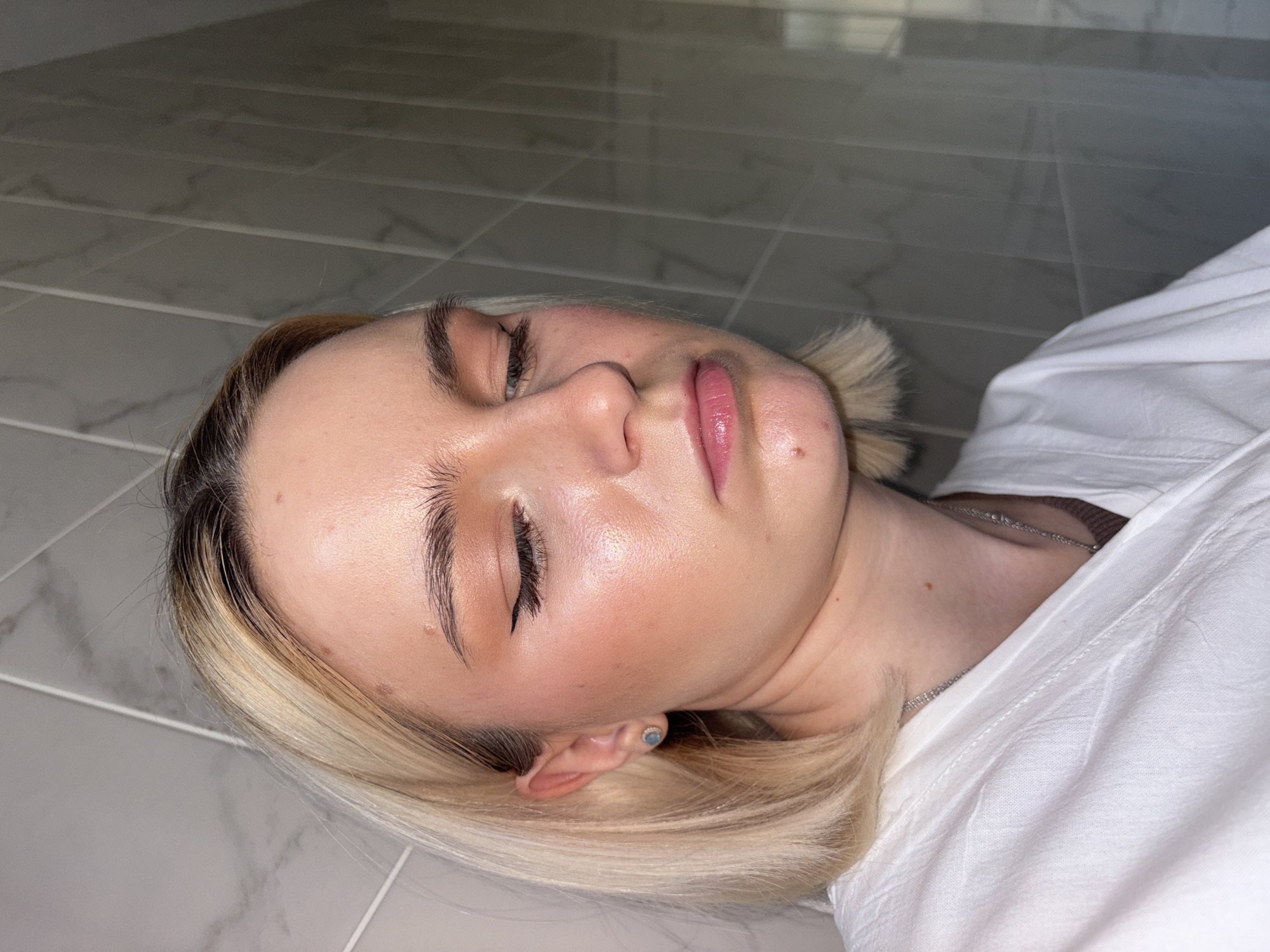 A young woman is depicted lying on a light grey tiled floor, her head resting on the surface, in a high-angle, close-up shot. Her eyes are closed, suggesting she is asleep or resting. She has short blonde hair with darker roots, styled to frame her face, and prominent, well-groomed eyebrows. Her face shows subtle makeup, including a pinkish lip gloss and blush that highlights her cheekbones. A small, light blue stud earring is visible in her left ear. She is dressed in a white, possibly cotton, garment, and a delicate silver chain necklace is visible around her neck.
The tiled floor features a light grey and white marbled pattern with distinct grout lines, indicating an indoor setting such as a bathroom or a modern room. The lighting is soft, creating gentle reflections on her skin, particularly her forehead and cheeks, giving her complexion a luminous quality. The image captures a moment of quiet repose or unconsciousness. While the context suggests Ladozhskoe Ozero, Russia, there are no specific visual cues within the image itself to confirm this geographical location. No text is visible.