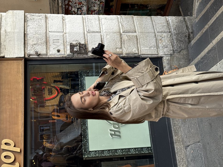 A young adult female stands outdoors on a paved surface in Venice, Italy, adjacent to the Hard Rock Cafe. She possesses long brown hair and is dressed in a beige trench coat over a plaid shirt. Her right hand holds a black compact digital camera, positioned facing her, while her left hand holds a dark-colored smartphone to her left ear, indicating an active phone call or audio engagement. Her gaze is directed slightly upwards and to the right, with her mouth slightly open. A silver-colored ring is visible on her right ring finger.
To her left, a large storefront window displays a red neon "HARD ROCK CAFE" sign and a light-colored acoustic guitar. Reflections of people are visible in the window glass. A wooden sign above the window partially shows "op," consistent with "Hard Rock Shop." To her right, a weathered building facade constructed from light-colored stone blocks features the Roman numeral "VII" carved into one block. Below this, an older intercom system is mounted on the wall, and further right, the wall segment is covered extensively with stickers and minor graffiti. The ground consists of dark-toned pavement with lighter horizontal stripes.
