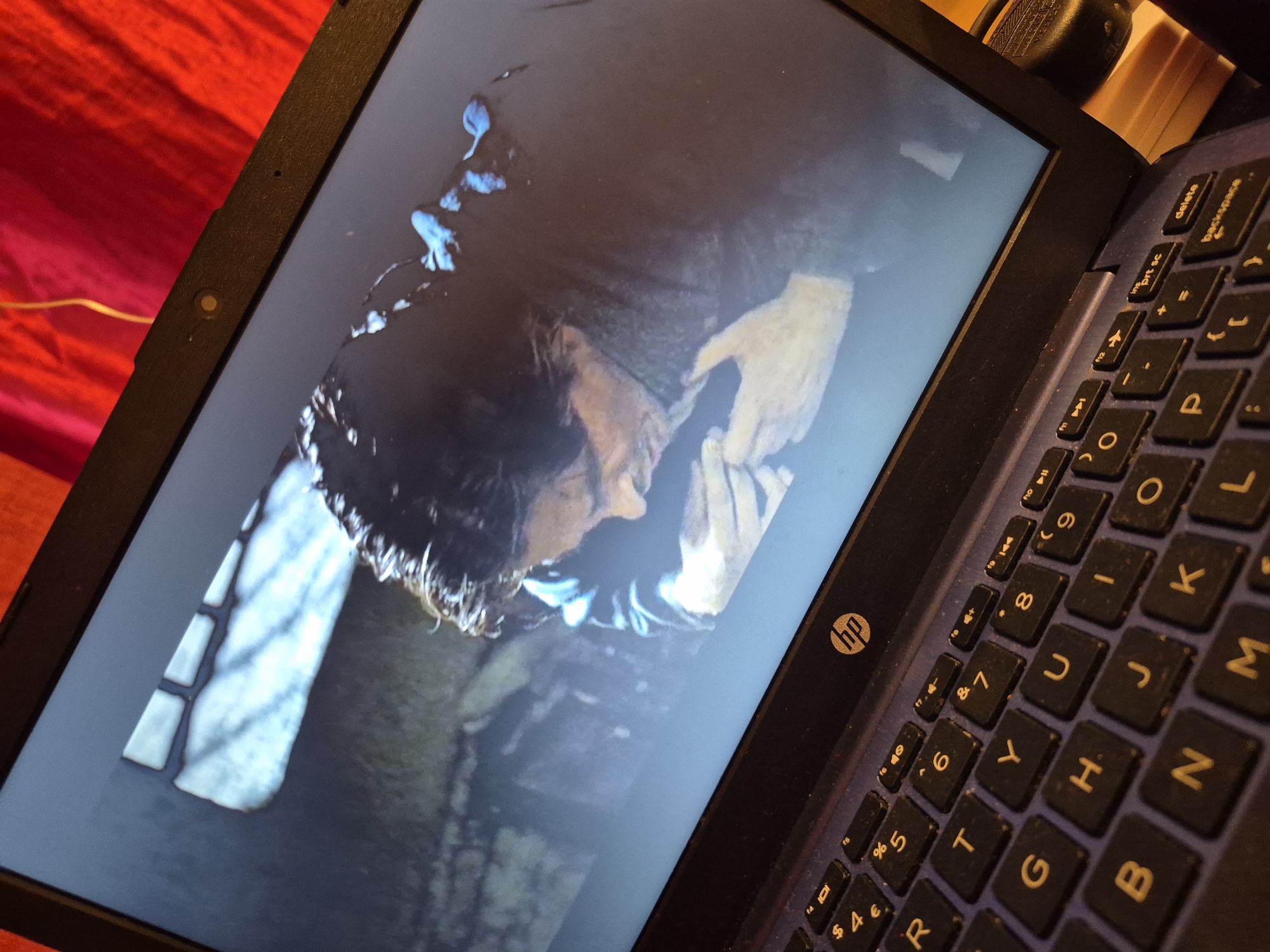 A laptop displays a dimly lit scene with a person hunched over, their face partially obscured. The screen shows a close-up of the person's head and shoulders, with a blurred background. The laptop itself is open, with a keyboard visible in the foreground. The keyboard is a dark color with lighter-colored keys, and some keys are illuminated by the screen's glow. The laptop is placed on a surface, and a red fabric is visible behind it, suggesting a domestic or indoor setting. No specific location in Sandwell, United Kingdom can be determined from this image.