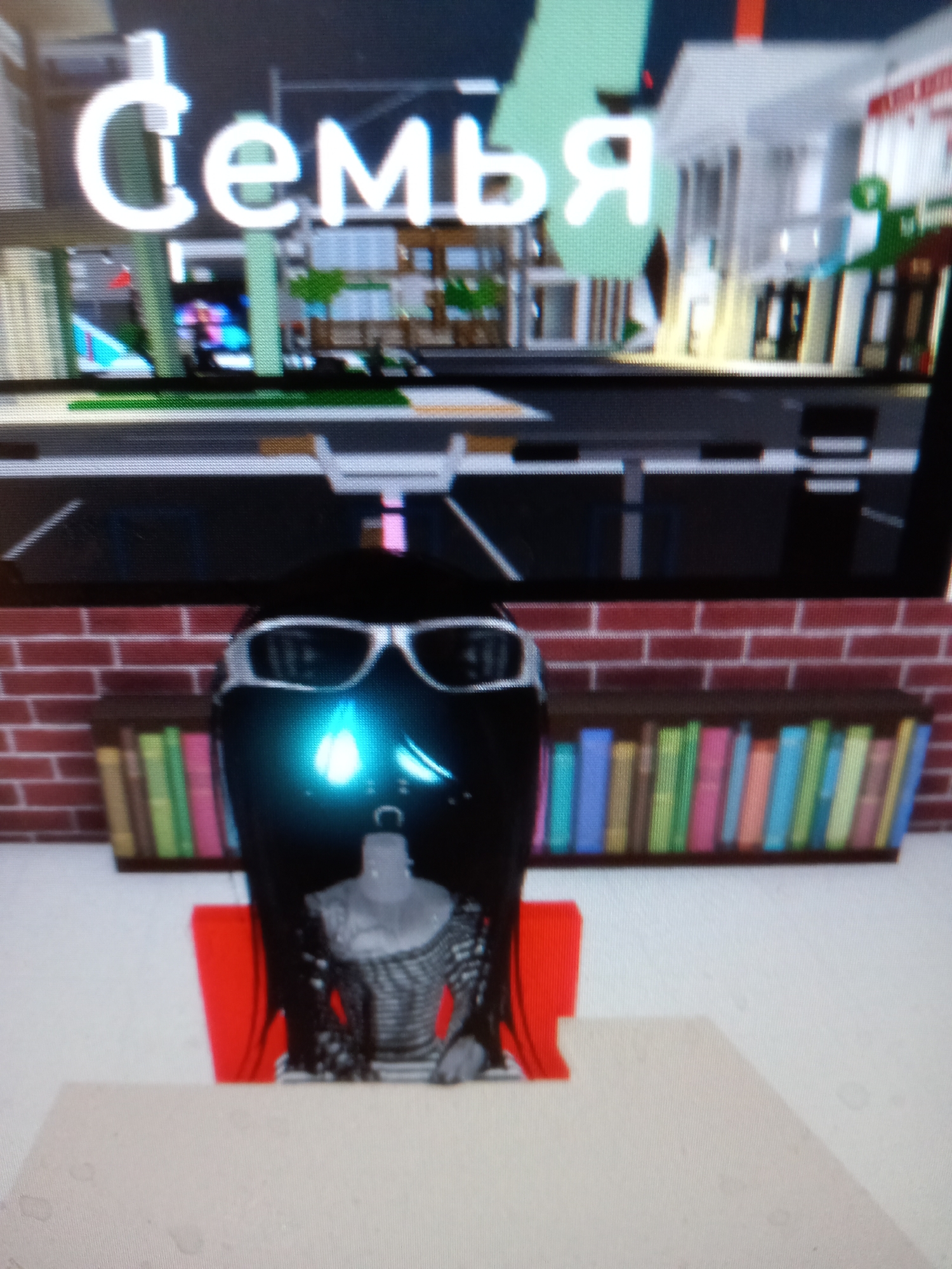 The image displays a screen showing a video game environment. At the top, large white Cyrillic letters spell out "Семья," which translates to "Family." The background appears to be a simulated city scene with buildings and trees. Below this, a black, helmet-like object with silver-rimmed sunglasses is positioned in the foreground. A light blue, flame-like glow emanates from within the helmet. Below the helmet, a mannequin torso wearing a black and white checkered patterned shirt is visible. Behind the helmet and mannequin, a brick wall serves as a backdrop for a bookshelf filled with colorful books in various shades of blue, green, and pink. A bright red object, possibly a chair or stand, is partially visible to the left of the mannequin. The image is taken from a slight angle, and the lighting suggests an indoor setting or a screen being viewed in a dimly lit room. There is no visible information in the image to suggest the location is the city of Rudny, Kazakhstan.