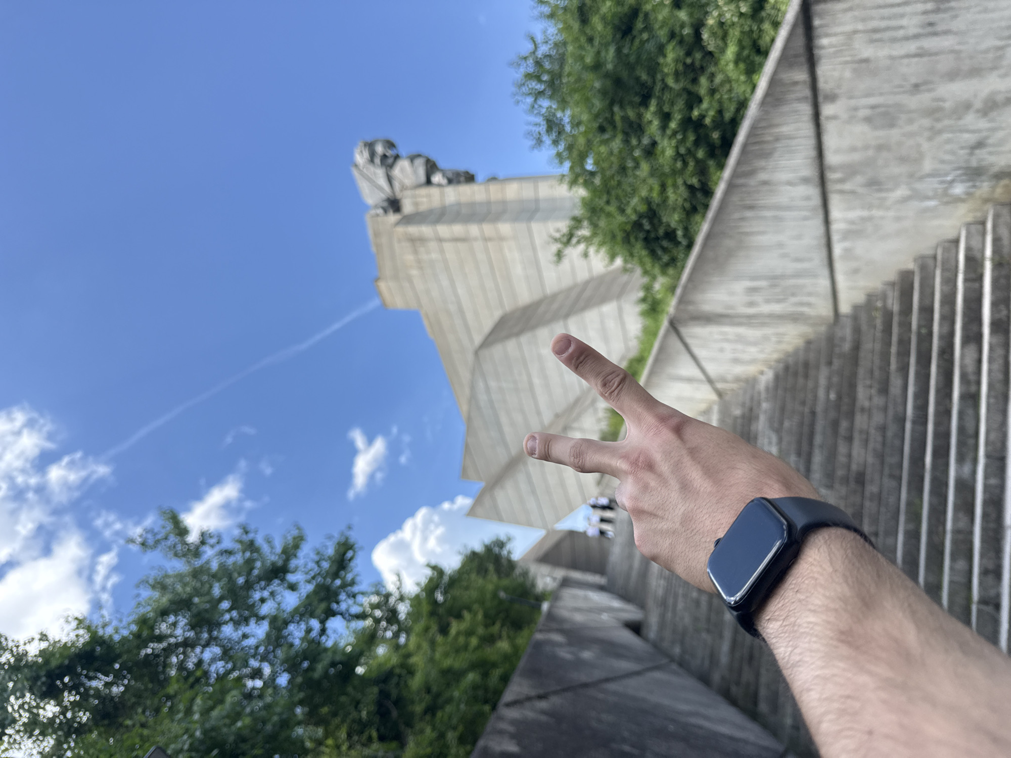 The image is a low-angle shot taken outdoors on a sunny day, featuring a person's arm and hand in the foreground. The hand is making a peace sign, with the index and middle fingers extended. The person is wearing a black smartwatch on their wrist.
In the background, there is a large concrete monument, which is part of the "Founders of the Bulgarian State" monument complex in Shumen, Bulgaria. The monument is set against a blue sky with a few clouds. There is also some greenery visible, including trees and shrubs. The photograph is taken from a slightly upward angle, suggesting the photographer is below the monument and possibly on a set of stairs leading up to it.