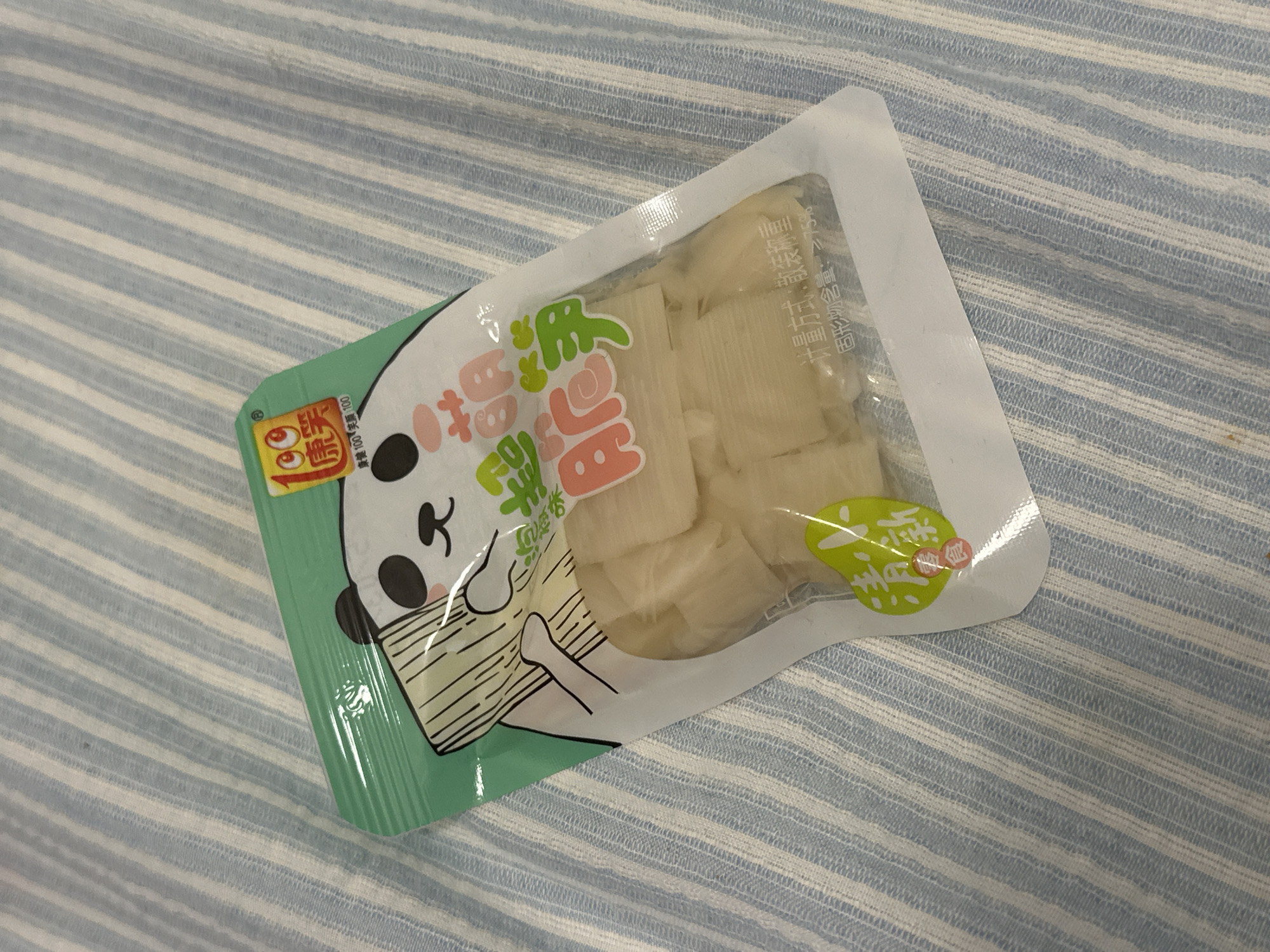 The image shows a package of pickled bamboo shoots. The package is mostly white and clear, with a green corner that features a cartoon panda design. The package contains pieces of light-colored, rectangular bamboo shoots. The packaging has some Chinese writing on it. The package rests on a light-blue striped fabric.
