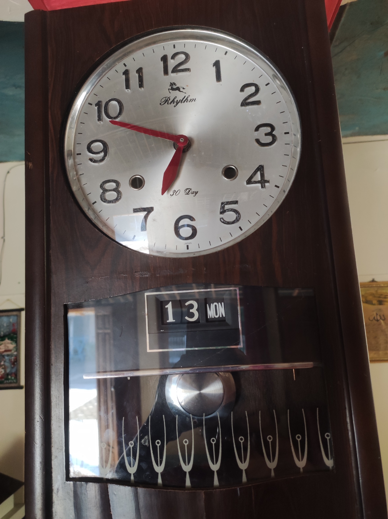 The image shows a close-up view of a vintage Rhythm wall clock, likely located in Mojopuro, Indonesia. The clock has a round, silver face with black hour markers and Roman numerals. The hands are red. The clock also features a date display showing "13 MON". Below the clock face, there's a decorative panel with a series of white, stylized designs that resemble tuning forks or musical notes. The clock is housed in a dark wooden casing. The background suggests an indoor setting with a wall and possibly a window or another object visible.