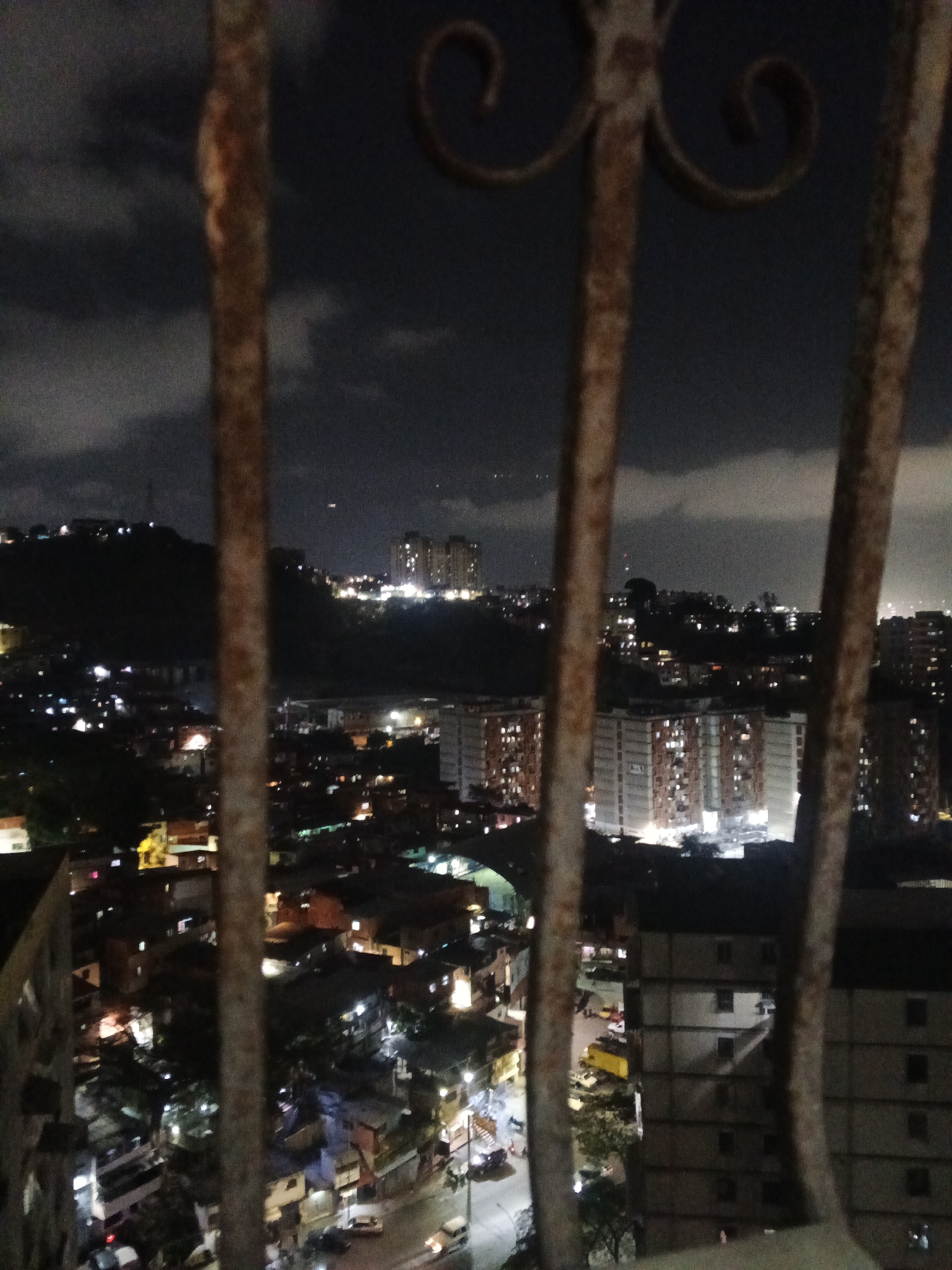 A nighttime view of Caracas, Venezuela, as seen through a rusty, ornate balcony railing. The cityscape is illuminated by scattered lights from buildings and street lamps. A hill covered with buildings is visible in the background, partially obscured by a cloudy night sky. In the foreground, residential and commercial buildings are interspersed with streets, where car and streetlight illumination can be seen. The photo is taken from a high vantage point, giving a perspective overlooking a densely populated area. The focus is slightly blurred, adding to the atmospheric quality of the night scene.