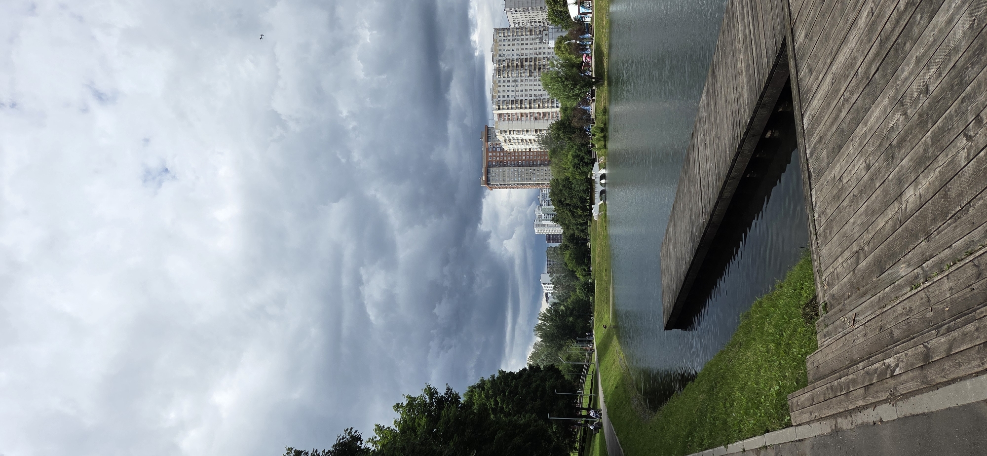 The image depicts a view of the Serebryany Bor park in Moscow, Russia, on a cloudy day. In the foreground, a wooden pier juts out into a calm body of water. The pier is constructed of weathered, aged planks. To the left of the pier, a grassy embankment slopes down to the water, bordered by a paved walkway. On the walkway, a few individuals are visible, one on a bicycle and others appearing to be seated or strolling. Trees with lush green foliage line the left side of the image, and further back, across the water, a picturesque park landscape is evident, featuring a white arched bridge and more trees. In the background, a cluster of modern high-rise apartment buildings forms the urban skyline, contrasting with the natural park setting. The sky is filled with heavy, dark gray clouds, suggesting impending rain or overcast weather. A small dark object, possibly a bird or drone, is visible in the upper right quadrant of the sky.