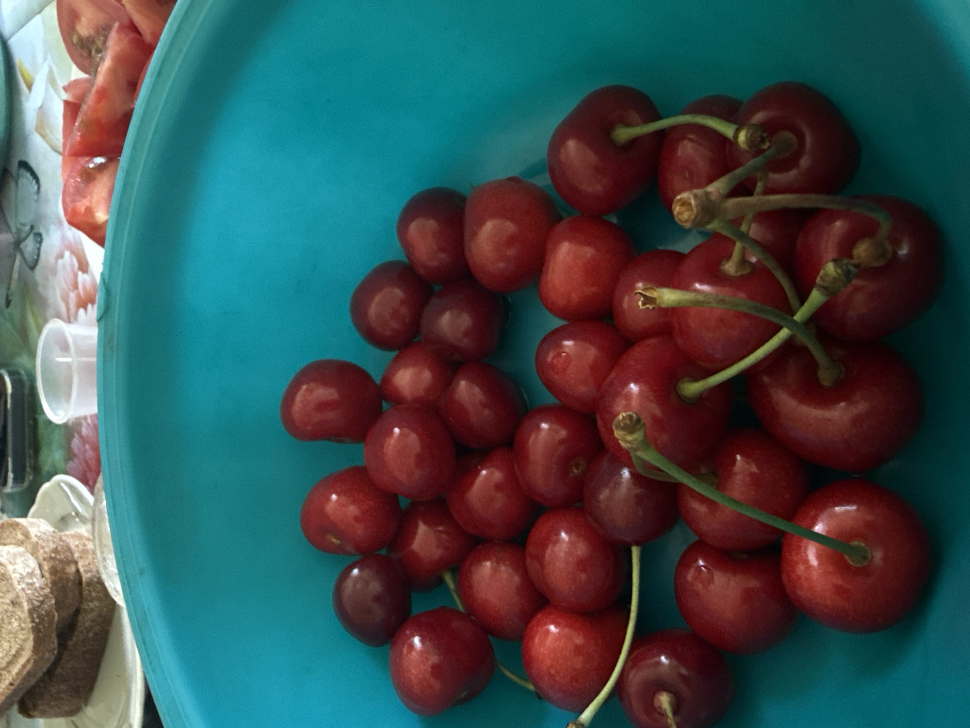 A close-up, overhead shot features a vibrant turquoise bowl overflowing with ripe, red cherries. The cherries are varied in size and shade, with some appearing darker and others a brighter red. Most of the cherries still have their green stems attached, some in clusters of two or three. The bowl itself is large and rounded, filling most of the frame. In the background, out of focus, are slices of bread and chopped tomatoes on a patterned tablecloth. There are no people visible in the image. The scene appears to be indoors, likely in a kitchen or dining area. There is no information to determine the location in Dnipro, Ukraine.