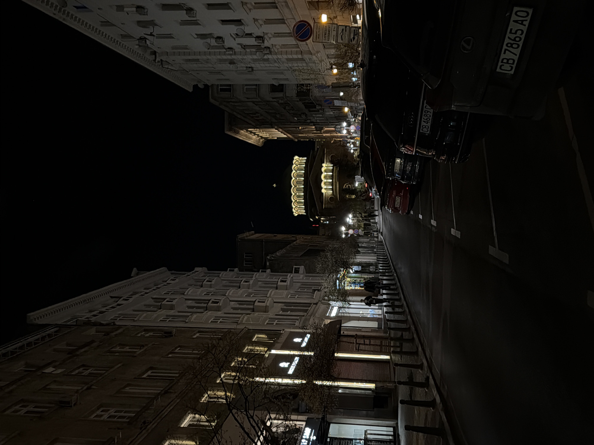 Here is a description of the image:
At night, a street in the city of Unknown, Unknown is captured in this image. The view is taken from street level, displaying buildings on either side and cars parked along the right edge. Above, the night sky is dark. The architectural styles are varied; some buildings are taller with multiple windows. Lights from the buildings and street lamps illuminate the scene. The road stretches into the distance. There are some people walking along the side of the road. A license plate on a parked car is visible.