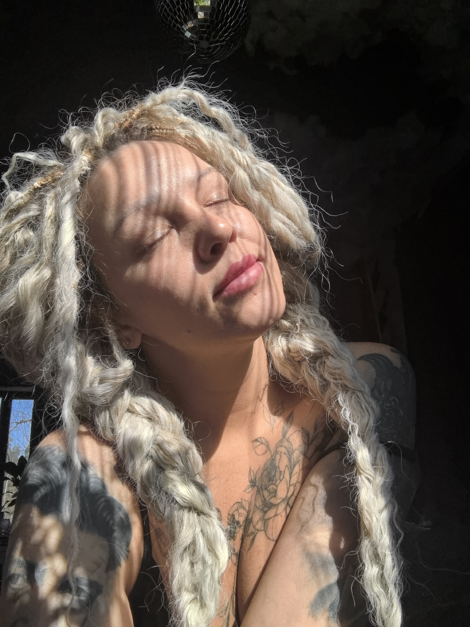 The image captures a person, likely female, indoors, basking in direct sunlight. Her eyes are closed, and a subtle smile plays on her lips, conveying a sense of peace and contentment as striped shadows fall across her face from an unseen obstruction, possibly window blinds. She has distinctive long, light-colored dreadlocks or heavily textured hair, which is a prominent feature, falling over her shoulders and chest.
The person is extensively tattooed, with intricate designs, including floral patterns, visible on her chest, shoulder, and arm. In the upper portion of the frame, a disco ball is partially visible, reflecting the bright sunlight. The background is largely dark, suggesting a dimly lit interior contrasting with the direct sun. Through an opening or window in the background, a glimpse of blue sky and tree branches hints at the outdoor setting.
The strong, directional light indicates it is daytime, likely a sunny morning or afternoon. The scene evokes a feeling of relaxation and warmth. There is no visible text in the image. This serene moment takes place indoors, potentially in Noviy, Russia, with the outdoors visible through a window.