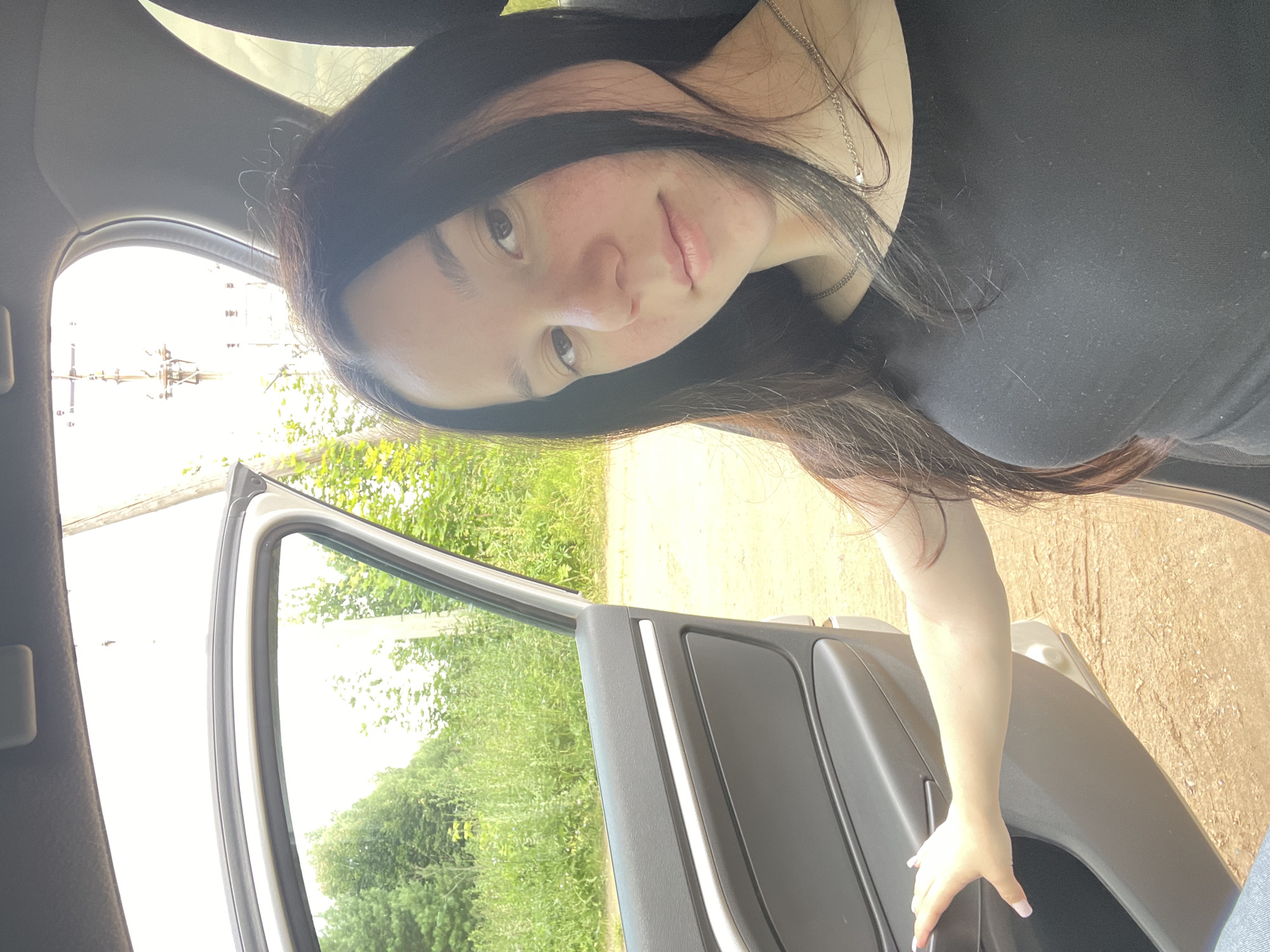 In this image, a young woman with long, dark hair and fair skin is captured in a candid moment inside a car. Her head is tilted, and she gazes towards the camera with a slight smile. She's wearing a black top and a delicate silver necklace.
The car door is open, revealing a backdrop of lush green trees and vegetation, suggesting an outdoor, possibly rural setting. The sunlight filtering through the leaves indicates it's daytime, likely a clear or partly cloudy day. The ground visible outside the car appears to be dirt or gravel.
The woman's hand is resting on the car door, her fingernails painted a light pink. The interior of the car, visible in the foreground, shows the dark color of the door panel. The overall impression is one of casualness and informality, perhaps a snapshot taken during a drive or a stop on a journey. There is no visible text in the image. The scene is set in Nozhovka, Russia.