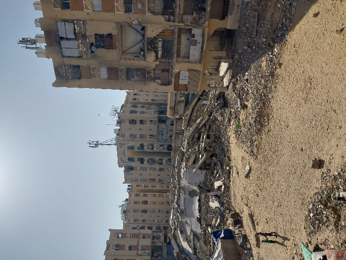 The image displays a devastated urban environment in Khan Yunis, Palestinian Territory, under a clear, bright sky with the sun visible in the upper-left. The scene is dominated by multiple multi-story residential buildings, primarily light brown in color, which exhibit extensive structural damage.
In the immediate foreground, a sandy, uneven expanse is littered with small pieces of concrete and debris. Two small figures, appearing to be children, are visible in the lower-left corner, standing or walking on the sandy ground, casting long shadows to their right.
Towards the mid-ground, substantial piles of rubble, twisted reinforcing bars, and fractured concrete indicate collapsed structures, filling the space between the foreground and the standing buildings. A blue tarpaulin is partially visible among the rubble on the left.
On the right side of the frame, a heavily damaged multi-story building stands partially intact. Its facade is severely scarred with numerous impact holes, blown-out windows, and exposed interiors, revealing structural elements. Further into the background, a row of similar apartment blocks extends across the horizon. These buildings also show considerable damage, including missing windows, pockmarked facades, and some areas appearing blackened. Many rooftops feature multiple communication antennas and other satellite or cellular equipment. One building in the center-background displays a distinctive arched decorative element on its upper facade. The overall environment is one of extensive destruction and desolation.