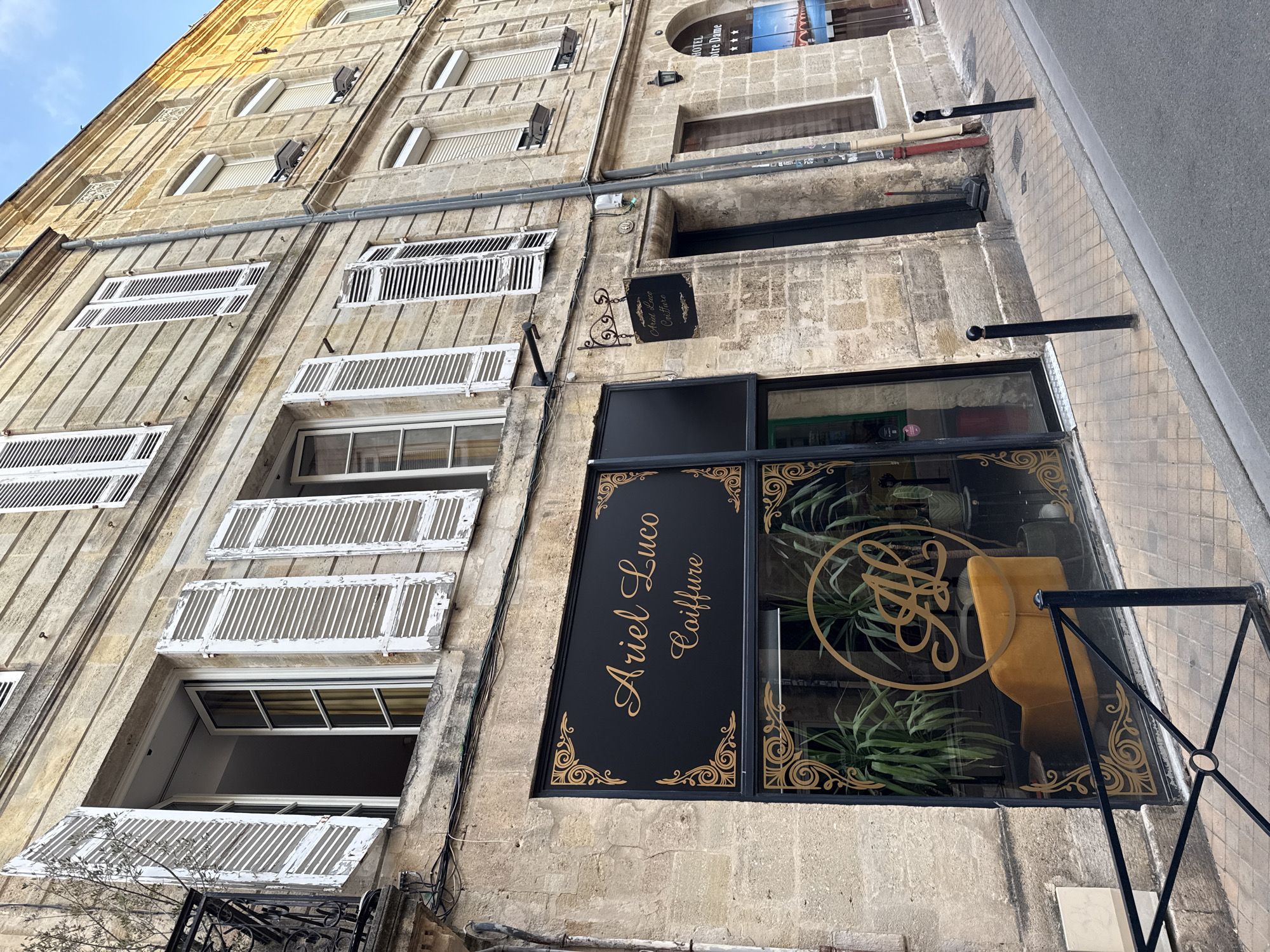 The image shows a street view in Bordeaux, France, featuring the facade of a building with a salon named "Ariel Luco Coiffure". The building is constructed of stone and has multiple windows with white shutters, some of which are open. The salon's storefront is prominent, with a black sign displaying the salon's name in gold lettering and decorative golden swirls. Through the window, a yellow armchair and plants are visible. A smaller sign hanging from the building also displays the salon's name. The street is paved with cobblestones, and there are bollards along the sidewalk. The sky is visible at the top of the image, indicating it is daytime. There are no people in the immediate visible area of the frame.