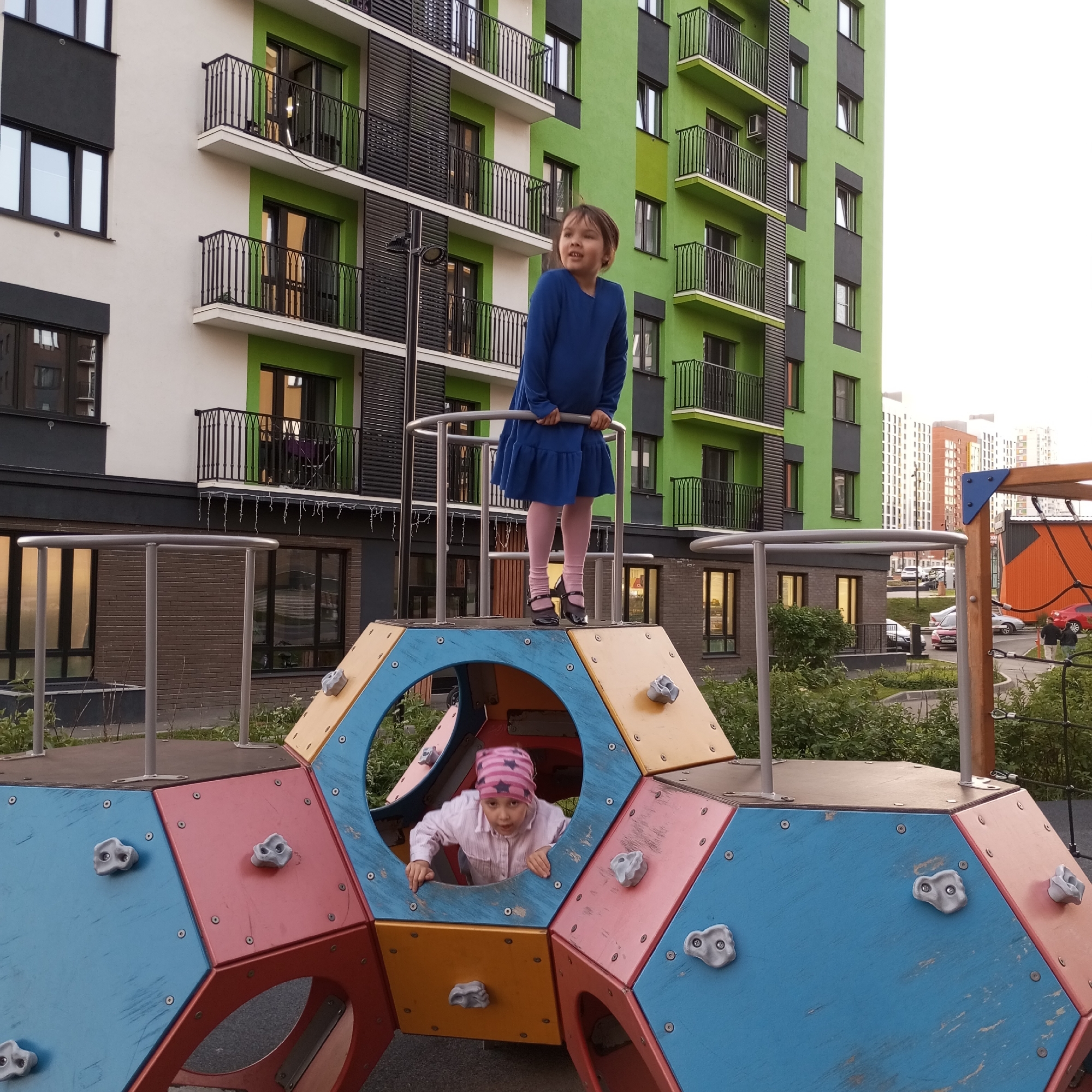 In Pervomaysky, Russia, a modern apartment complex with a distinctive green facade serves as the backdrop for a children's playground. Two young girls are engaged in play on the colorful, multi-faceted structure. One girl, wearing a blue dress and pink tights, stands atop the hexagonal play area, holding onto a metal railing. Her gaze is directed upwards and to her left. The second girl, clad in a pink-striped hat and striped shirt, is peeking out from a circular opening within the playground structure, her hands resting on the edge of the opening. The playground features climbing holds and is situated on a paved surface surrounded by greenery. The overall scene depicts a lively moment of childhood recreation in an urban setting.