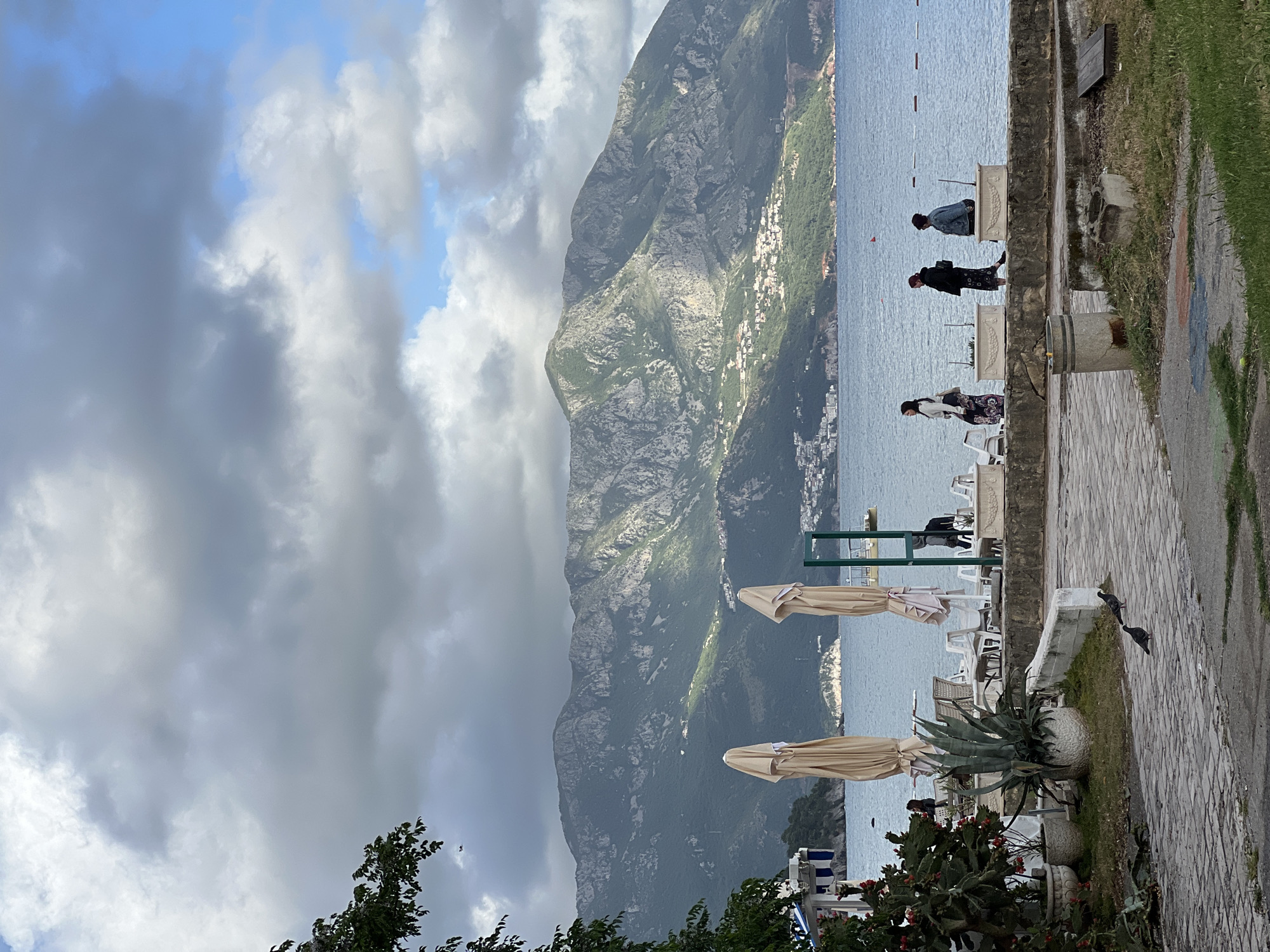 The image shows a view from a terrace overlooking the Adriatic Sea in Budva, Montenegro. Three people are standing along a stone ledge: a woman in a black and white patterned skirt, a person in a black jacket, and a person in a blue denim jacket. A large mountain dominates the background with buildings visible on its slope. The sky is overcast with grey and white clouds. Two closed parasols and white plastic furniture are seen on the terrace. Potted plants, including an agave, and a paved path are in the foreground. Two birds are on the path to the right.