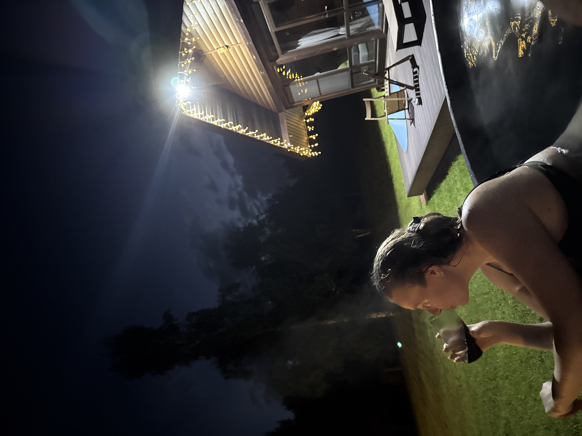 The scene depicts an outdoor nighttime setting, identified as being in the city of Копнино, Russia. A single female individual is centrally positioned in a dark-colored hot tub, observed from the mid-chest up. She possesses wet, dark hair and is wearing a dark garment consistent with swimwear. Her head is oriented to the left as she holds a clear glass containing a dark red liquid, likely wine, to her mouth in the act of consumption. Steam is visibly rising from the hot tub's surface, and the water reflects artificial light sources.
Adjacent to the hot tub is a raised wooden deck on which two wooden folding chairs and a small wooden table are arranged. Behind the deck stands a wooden house featuring a prominent gabled roof. The house displays a large window or glass door on its visible facade. Multiple strands of yellow string lights adorn the roofline and eaves of the house. A bright, artificial light fixture, possibly a floodlight, is mounted near the apex of the house, generating significant illumination and a distinct lens flare.
The immediate ground surrounding the hot tub and deck is covered in green grass, illuminated by the artificial light. Beyond the house and grass, the background consists of the dark silhouettes of trees under a night sky exhibiting some cloud cover. The scene is primarily illuminated by the artificial lights from the house, creating contrasts between brightly lit areas and deep shadows.
