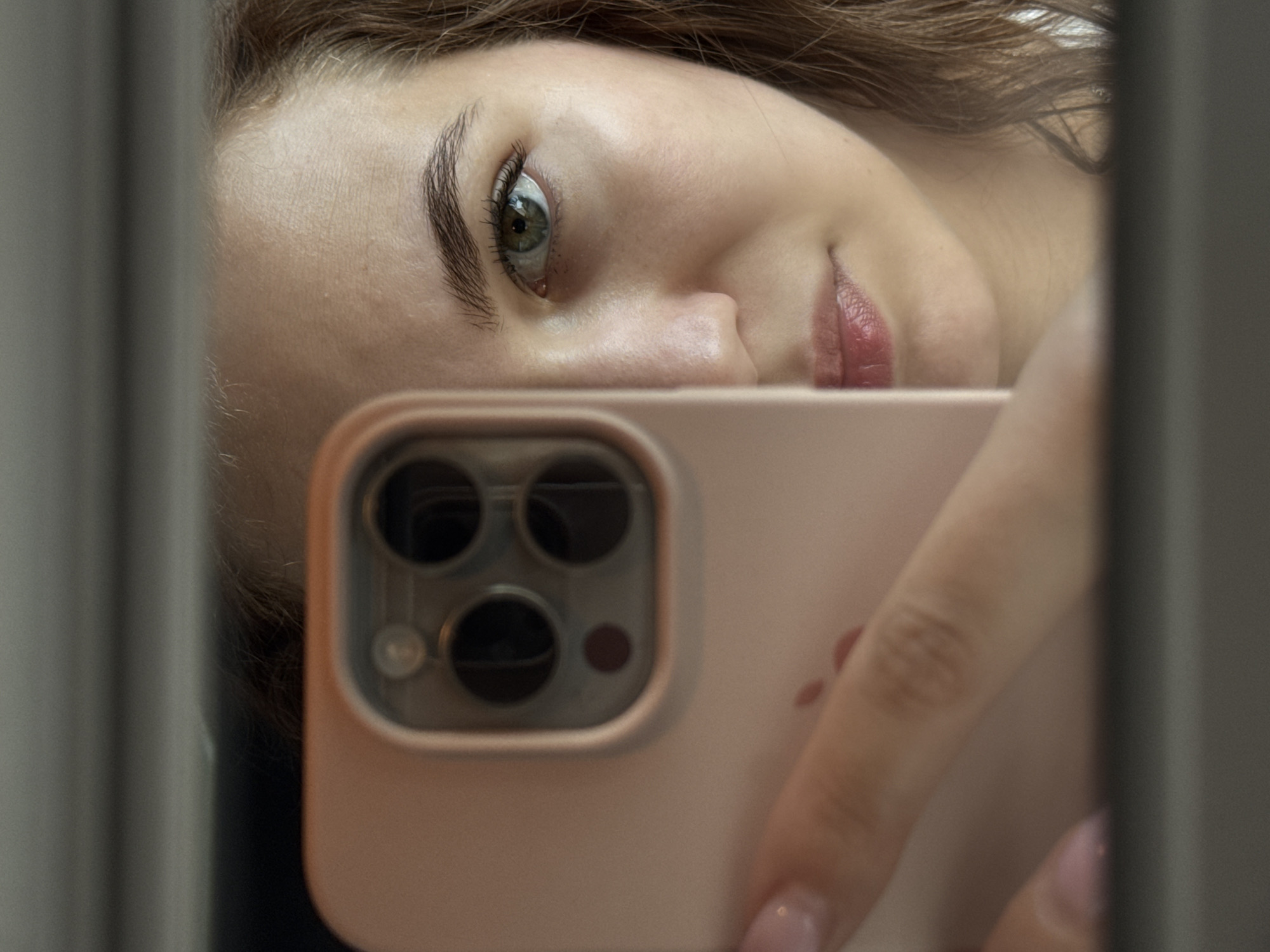 A selfie shot is reflected in a mirror. The subject, a light-skinned woman with brown hair, is positioned with her face angled downwards towards the camera. Her left eye is visible, and she has dark eyebrows and glossy lips. In the foreground, a pink smartphone partially obscures her face. The phone's camera lens and flash are visible. A hand with painted nails is holding the phone. The background reveals a blurry gray surface, possibly the mirror's edge or a wall. There is no verifiable information regarding the location of the scene being the city of Chaikovsky, Russia.