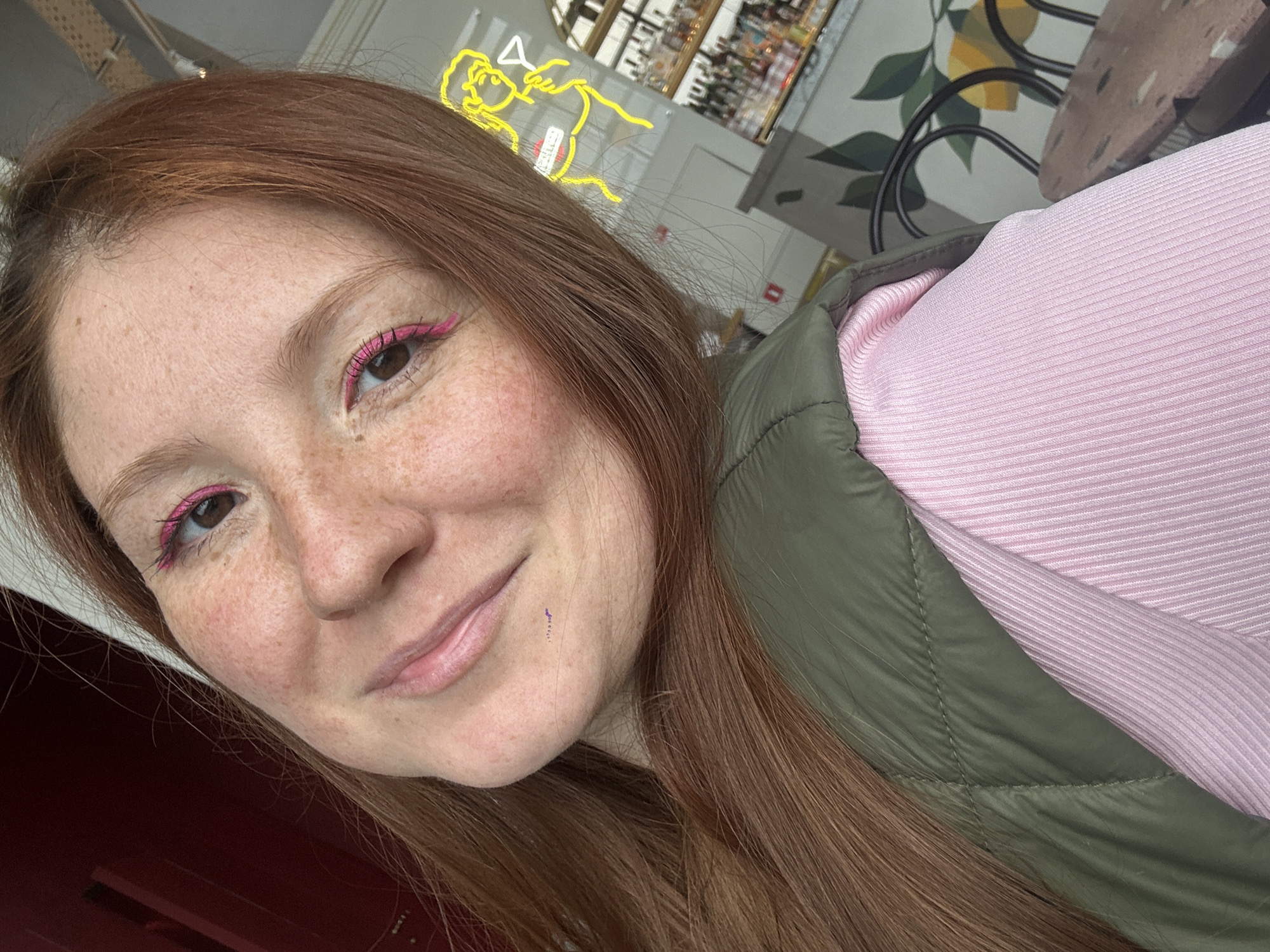 A selfie captures a woman with auburn hair and fair skin, marked with freckles, wearing a pink ribbed top and an olive green quilted vest. She has pink eyeliner applied. A vertical purple line is visible on her cheek.
In the background, a partially visible neon sign of two figures is visible. A bottle-lined bar is also visible behind her. A chair with lemon-patterned upholstery is also shown. The presence of the bar indicates that this picture was taken at a cafe, in Nizhny Novgorod, Russia.