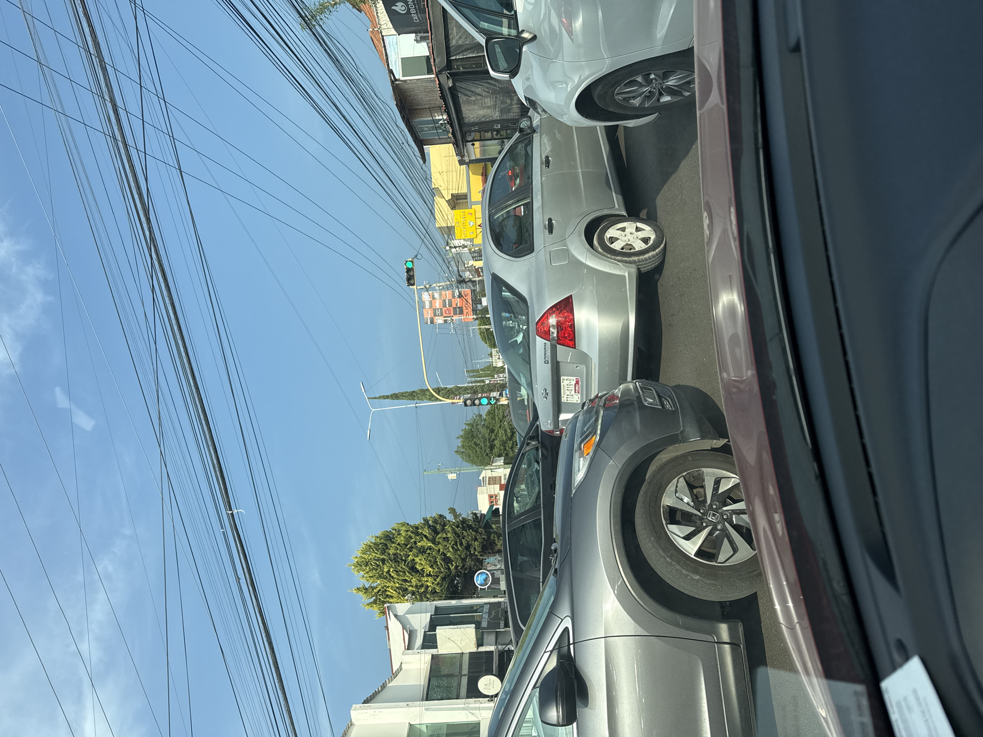 The image captures a vibrant urban street scene from the interior of a vehicle, looking out onto a bright, sunny day. The perspective is sideways, suggesting the original photo might have been taken with a phone held horizontally.
Numerous black power and communication lines crisscross the clear, bright blue sky, dominating the upper portion of the frame. Below these wires, a busy street is visible, populated by several cars, mostly silver and grey in color, either parked or in traffic.
In the foreground, the side and front wheel of a dark grey vehicle are prominent, hinting at the observer's position. Further along, a silver sedan displays the word "PERICANA" on its rear and a partially visible license plate reading "TTA-419-B". Another silver car is directly behind it.
Along the street, various buildings are visible, including one with a dark awning and a sign displaying "CARBONARIA". Another building appears yellow. A green traffic light in the distance signals that traffic is flowing. Green trees and other foliage punctuate the urban landscape. The overall impression is a typical daytime scene in a city, with clear weather and no discernible human activity beyond the presence of vehicles. The visual cues, including the license plate format and the general street environment, align with the provided context of Puebla, Mexico.