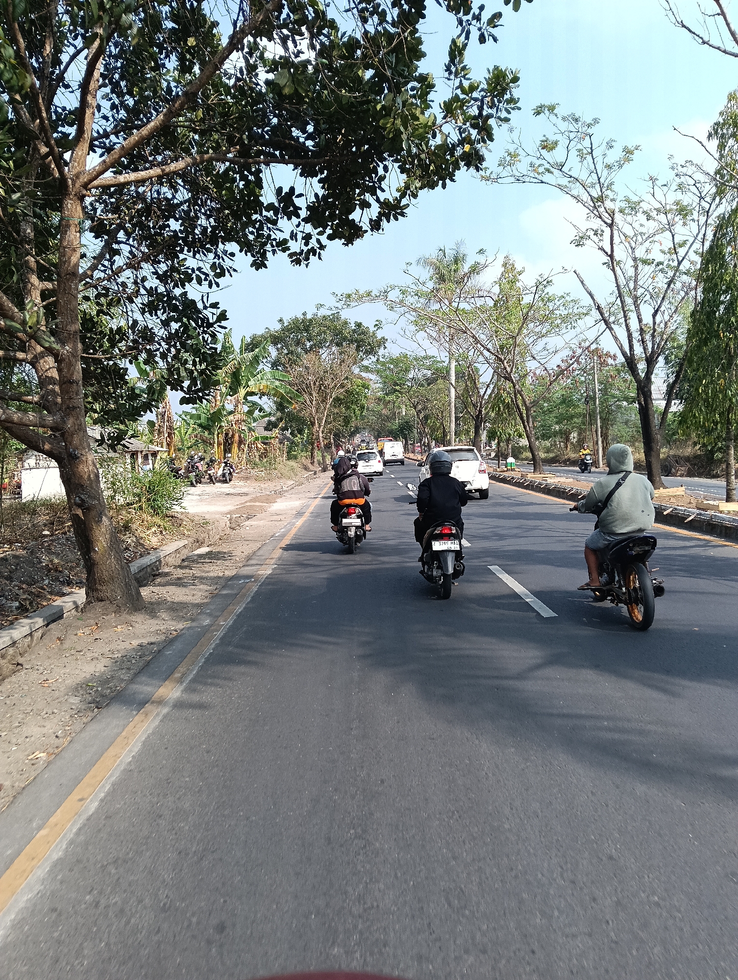 The image depicts a street scene in Tasikmalaya, Indonesia, under a clear blue sky. The foreground shows a paved road with a yellow line marking its edge and white lines indicating lanes. Several motorcycles are on the road. In the mid-ground, a white car is traveling in the same direction as the motorcycles. To the left of the road, there is a dirt area with some vegetation and a few parked motorcycles. Trees line the sides of the road, with foliage ranging from lush green to bare branches, suggesting a tropical or subtropical climate. The overall impression is that of a typical day in a city with active road traffic and natural elements integrated into the urban landscape.