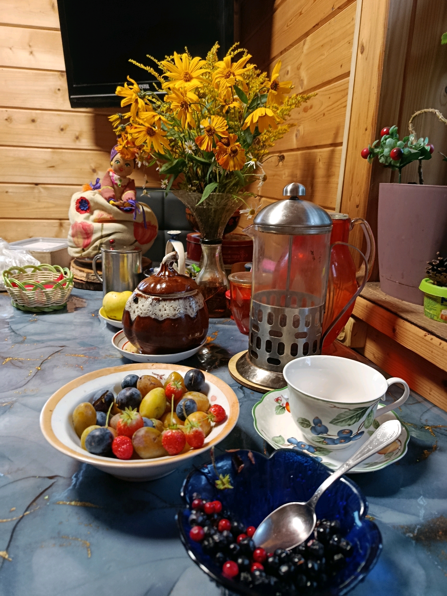 In an indoor setting with wooden paneled walls, a rustic wooden table is laden with various items. A bouquet of yellow flowers is prominent, sitting in a clear vase next to a decorative doll. In front of the flowers is a metal and glass French press, with a red pitcher to its right. To the left of the flowers is a metal mug and a small woven basket. Further to the right, a pink pot holds a small green plant with red berries.
On the table, a brown ceramic pot with a white glaze sits on a saucer. Next to it, a yellow apple rests on its own saucer. A plate in the foreground contains a medley of fruits including plums, strawberries, and grapes. To the right, a teacup with a blueberry pattern sits on a saucer, with a spoon placed inside a blue, heart-shaped bowl filled with blueberries and red berries. The overall impression is a still life arrangement, possibly depicting a tea or dessert setting.