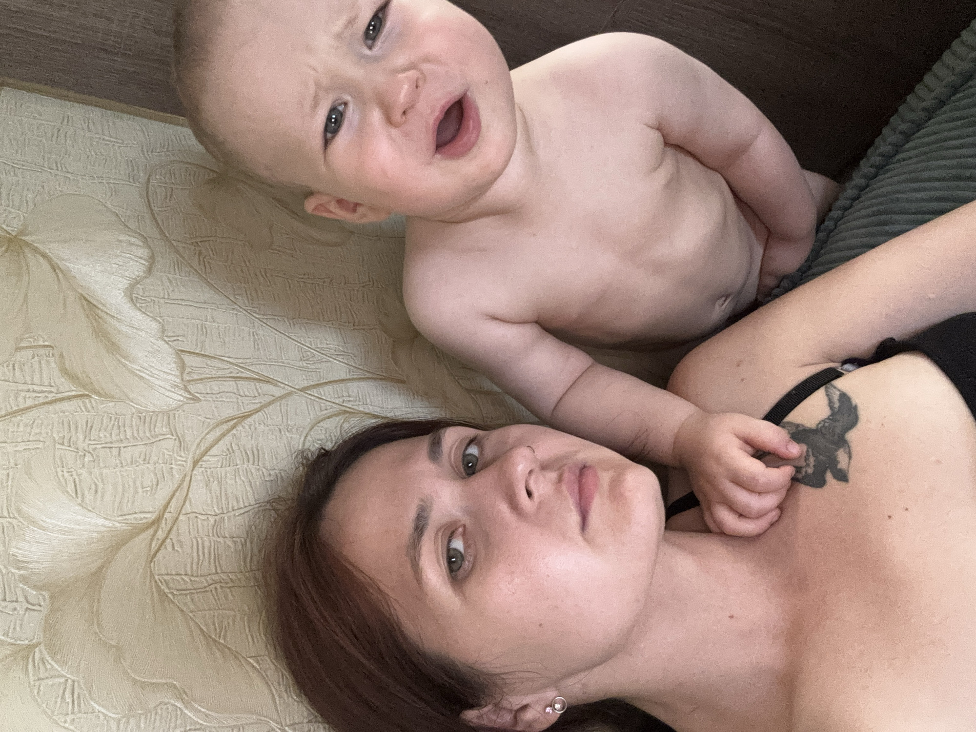 The media file shows a close-up, overhead shot of a woman and a baby lying down together. The woman is in the foreground, looking up at the camera with a neutral expression, her head tilted to her left. She has brown hair and is wearing a black top, with a tattoo of a moth visible on her right shoulder. The baby is positioned behind her, looking upwards with a surprised or questioning expression and mouth slightly open. The baby appears to be very young, with fair skin and grey eyes. Both subjects are lying on a textured surface that appears to be a bed or couch with a light beige, patterned sheet. The background is a darker, possibly brown or green, surface, which could be another part of the furniture. The lighting suggests it is daytime, likely indoors. No text is visible in the image. The overall scene depicts a moment of shared rest or play between a mother and her child.