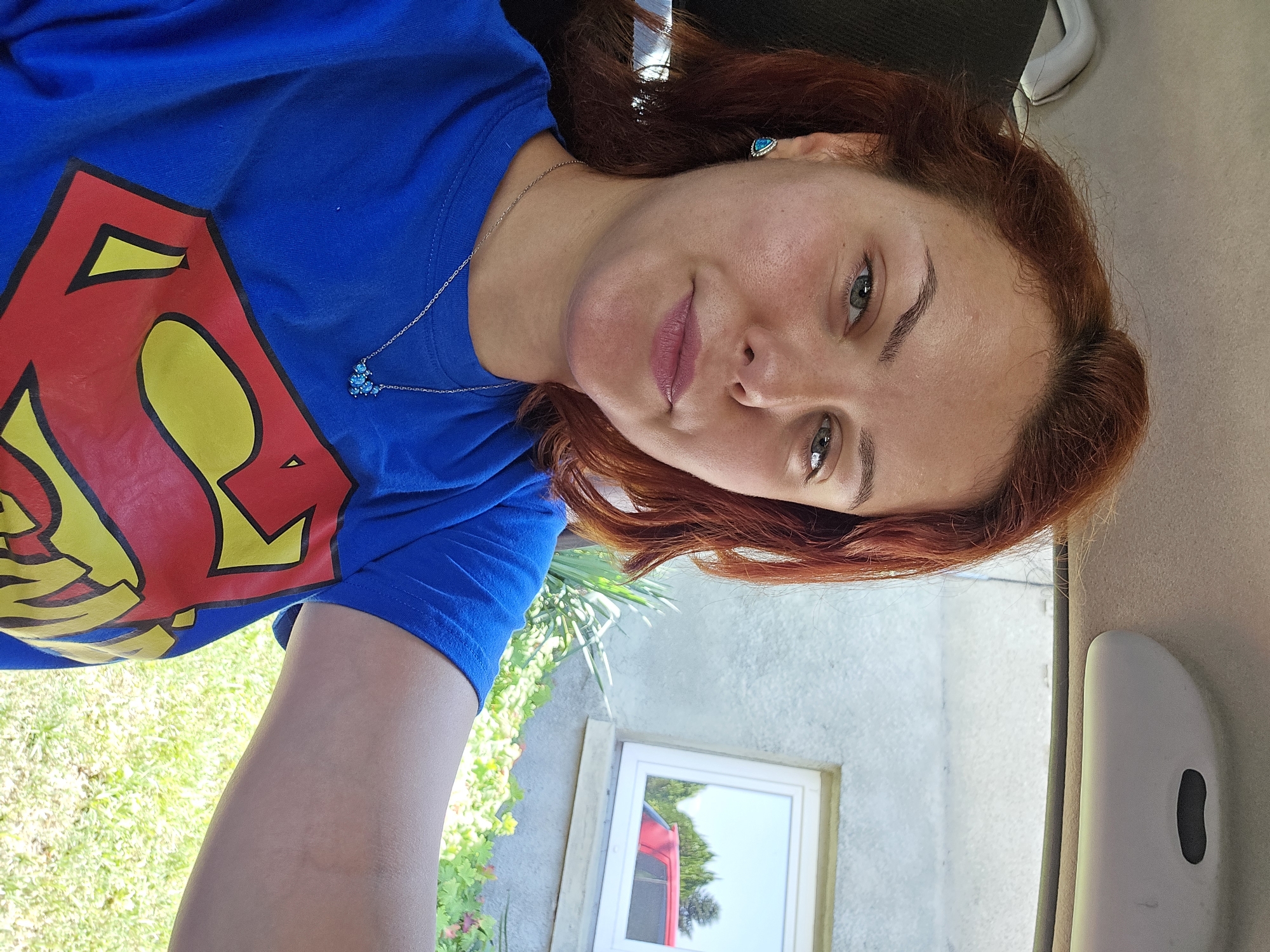 A woman with reddish-brown hair is captured in a selfie taken from inside a vehicle, as indicated by the car's interior visible in the upper portion of the image. She is wearing a blue t-shirt featuring the Superman logo. She has green eyes and is wearing a turquoise earring and a silver necklace with a cluster of blue beads. The background, visible through the car window, shows a grey building with a window, green foliage, and a red car reflected in the window. The overall setting suggests a casual, outdoor environment, possibly a residential area in Sofia, Bulgaria.