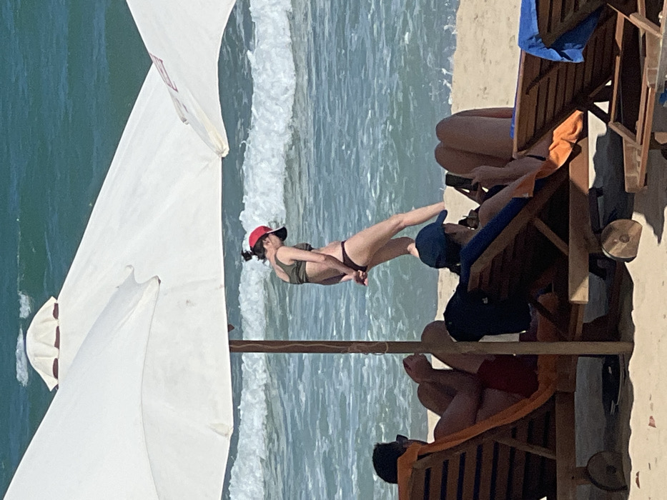 A woman wearing a red cap and a bikini lies on the beach near the water. There is a beach umbrella to her left. Several people are sitting in chairs nearby. The chairs and people are on a sandy beach close to the ocean.